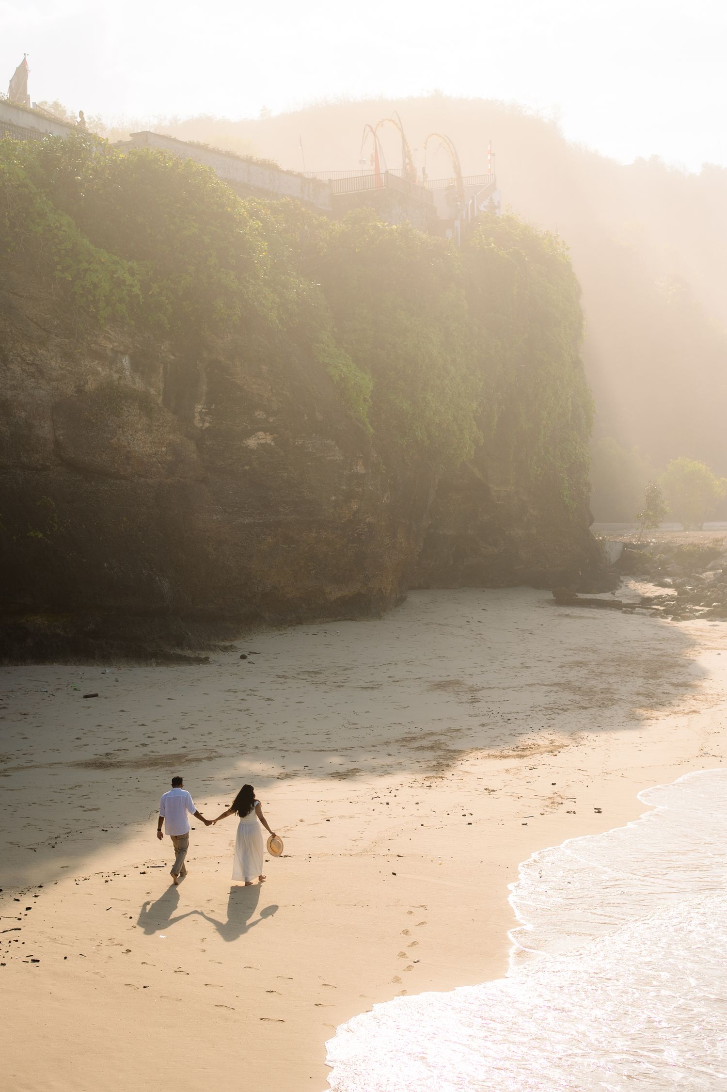 Couple holding hands walking on a misty beach at golden hour with dramatic cliffs in the background.