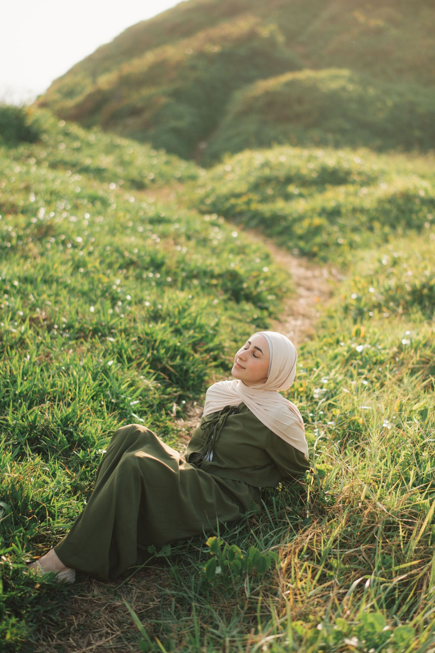 Woman in green hijab and dress sitting peacefully on grassy hillside path, eyes closed, enjoying warm sunlight.