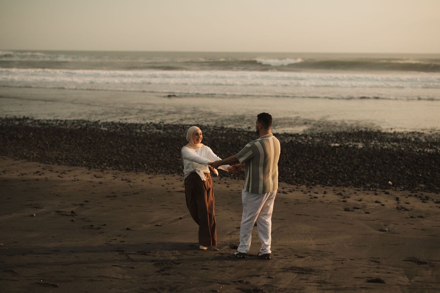 Couple holding hands on a dark sandy beach at sunset with ocean waves in the background.