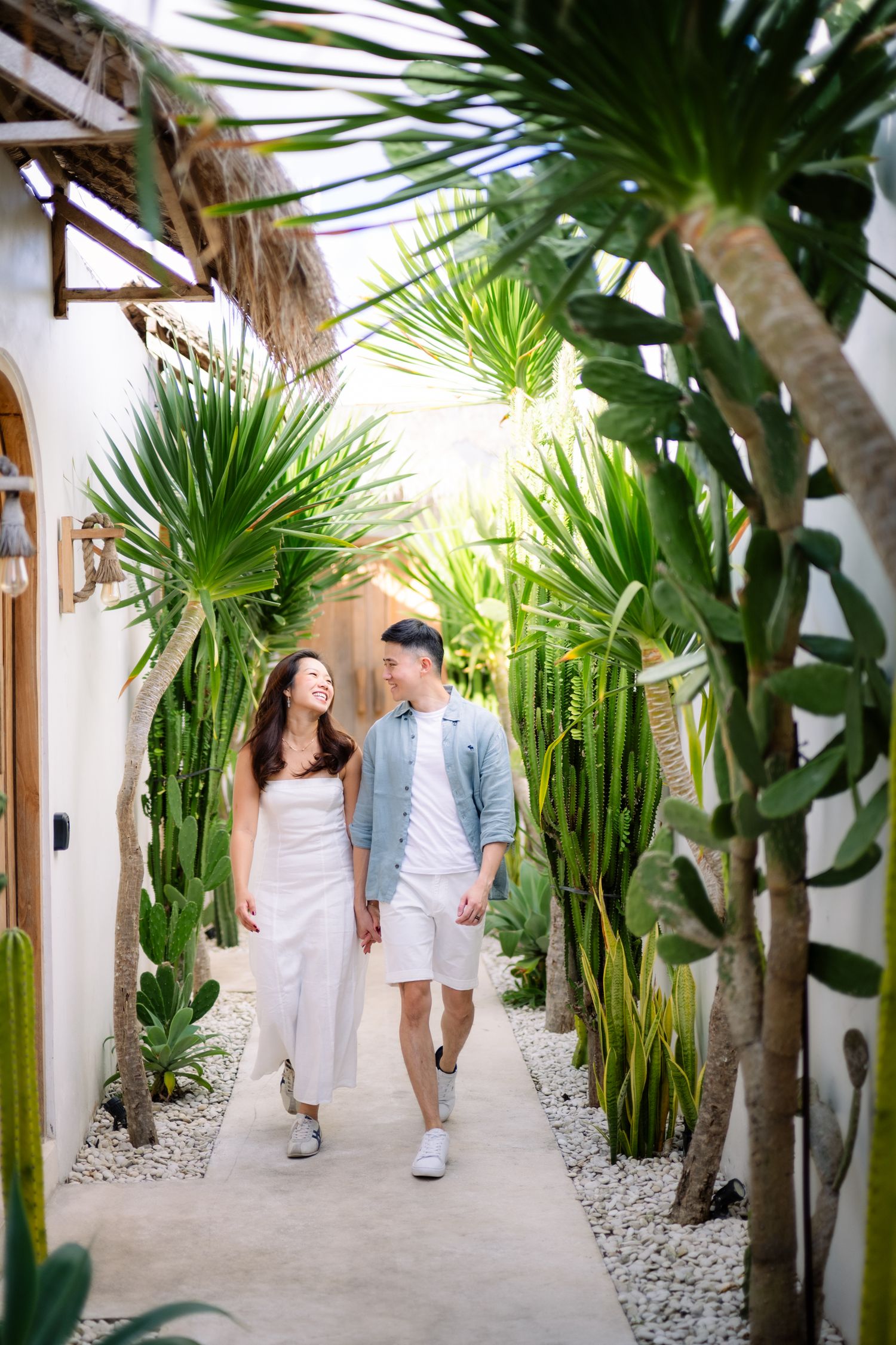 Couple walking hand-in-hand through a tropical garden pathway lined with cacti and palm trees near a white villa.