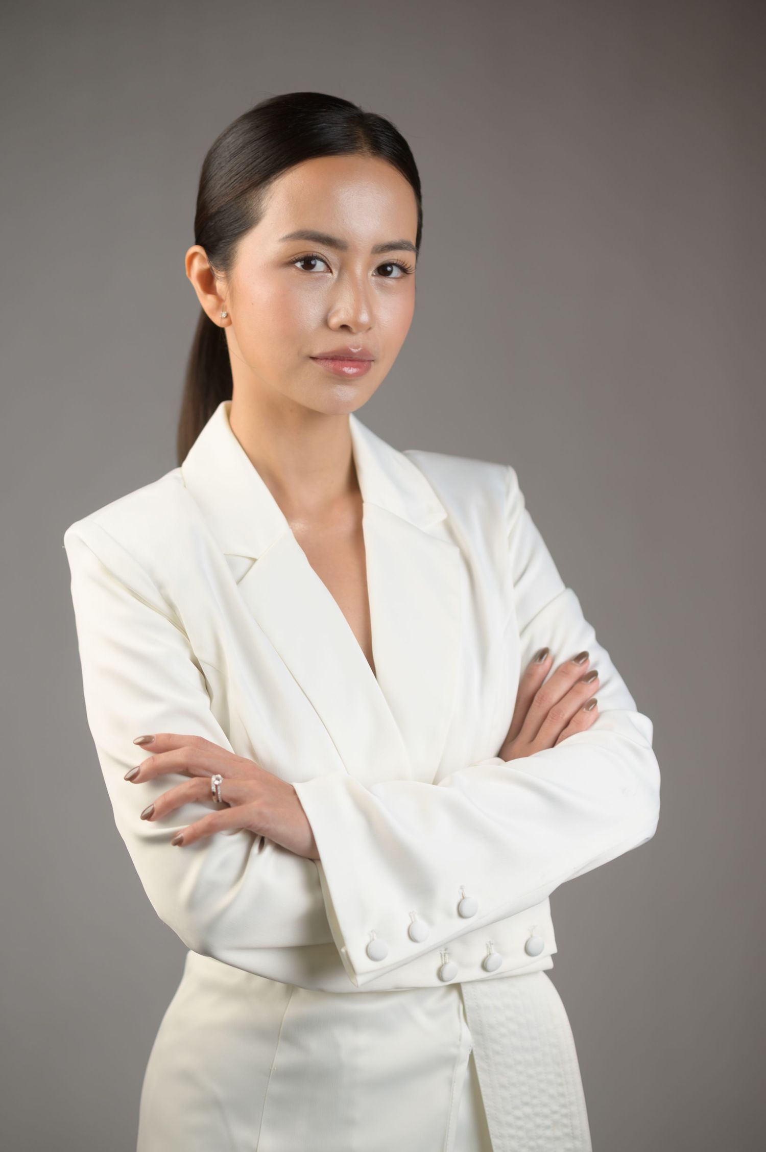 Professional Asian woman in white blazer with arms crossed, confident pose against gray background.