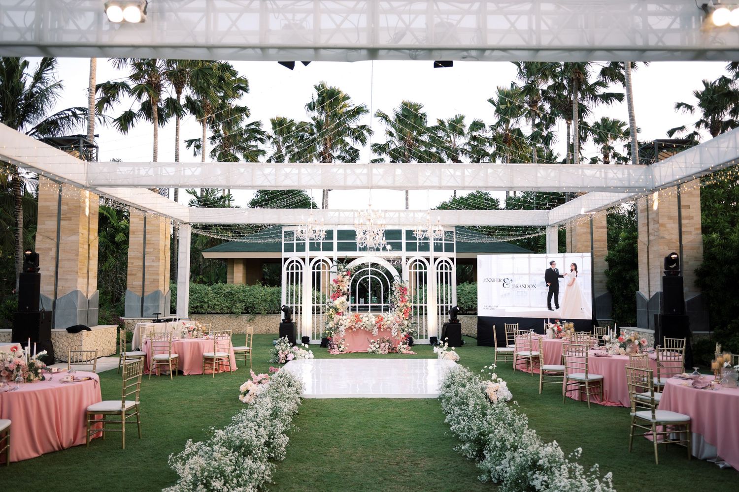 Elegant outdoor wedding reception with pink tablecloths, floral arch, palm trees, and string lights on manicured lawn.
