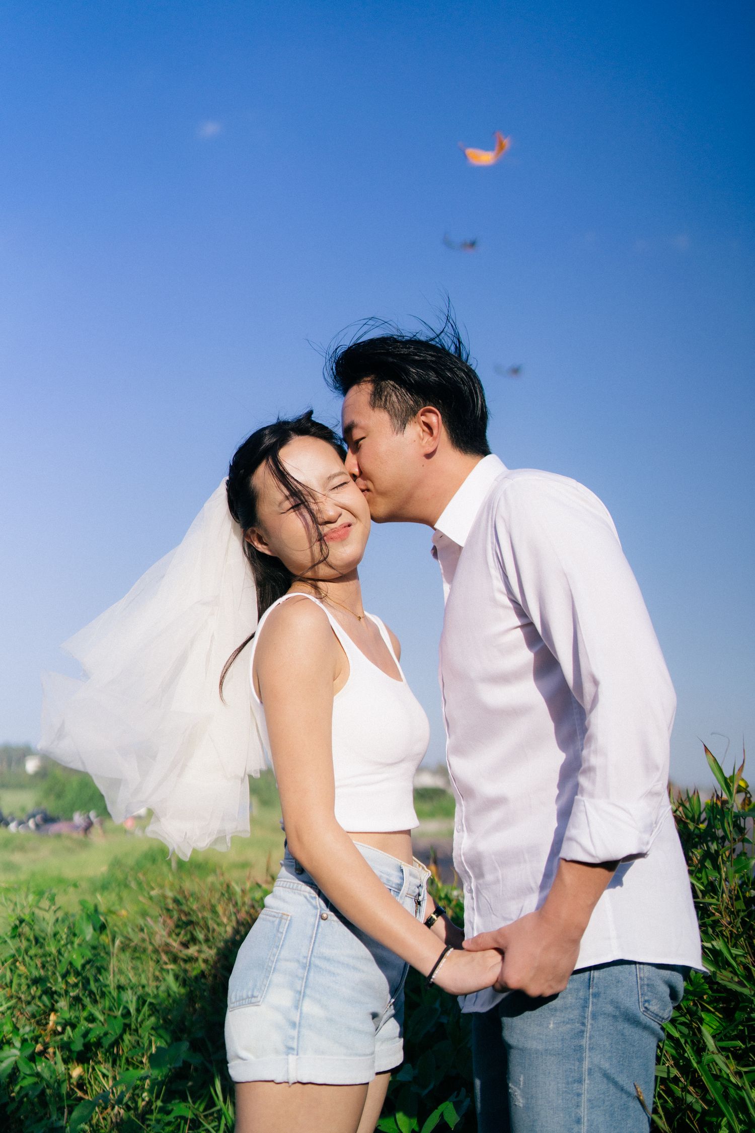 A couple shares a romantic kiss outdoors, bride in white crop top and veil, groom in white shirt, holding hands under blue sky.