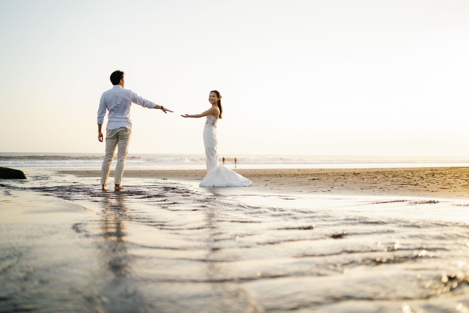 A bride and groom hold hands on a serene beach at sunset, with waves gently washing around their feet.