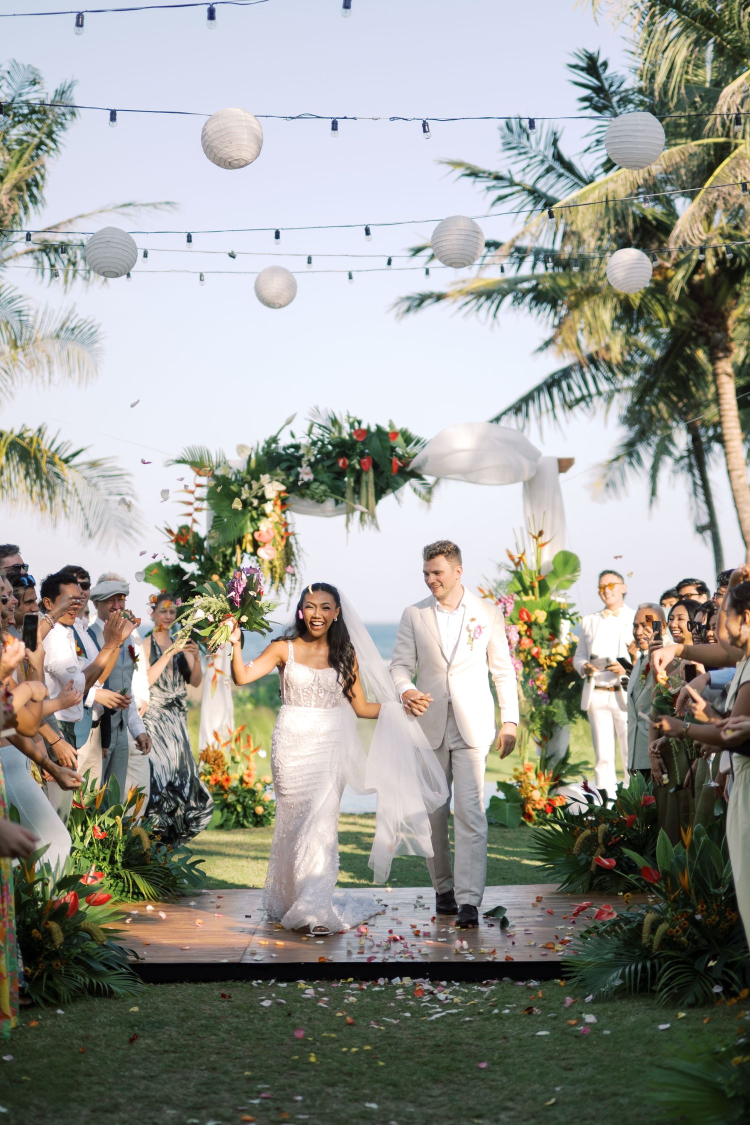 Bride and groom walk down tropical outdoor aisle with palm trees, floral arch, lanterns, and confetti at beach wedding.