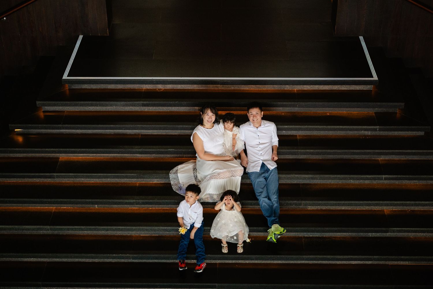 Family of four posing on wooden steps, parents seated with two young children in a warmly lit indoor staircase.
