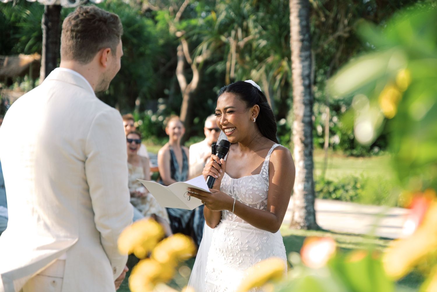 Bride in lace gown reads vows while smiling at groom during outdoor tropical ceremony.