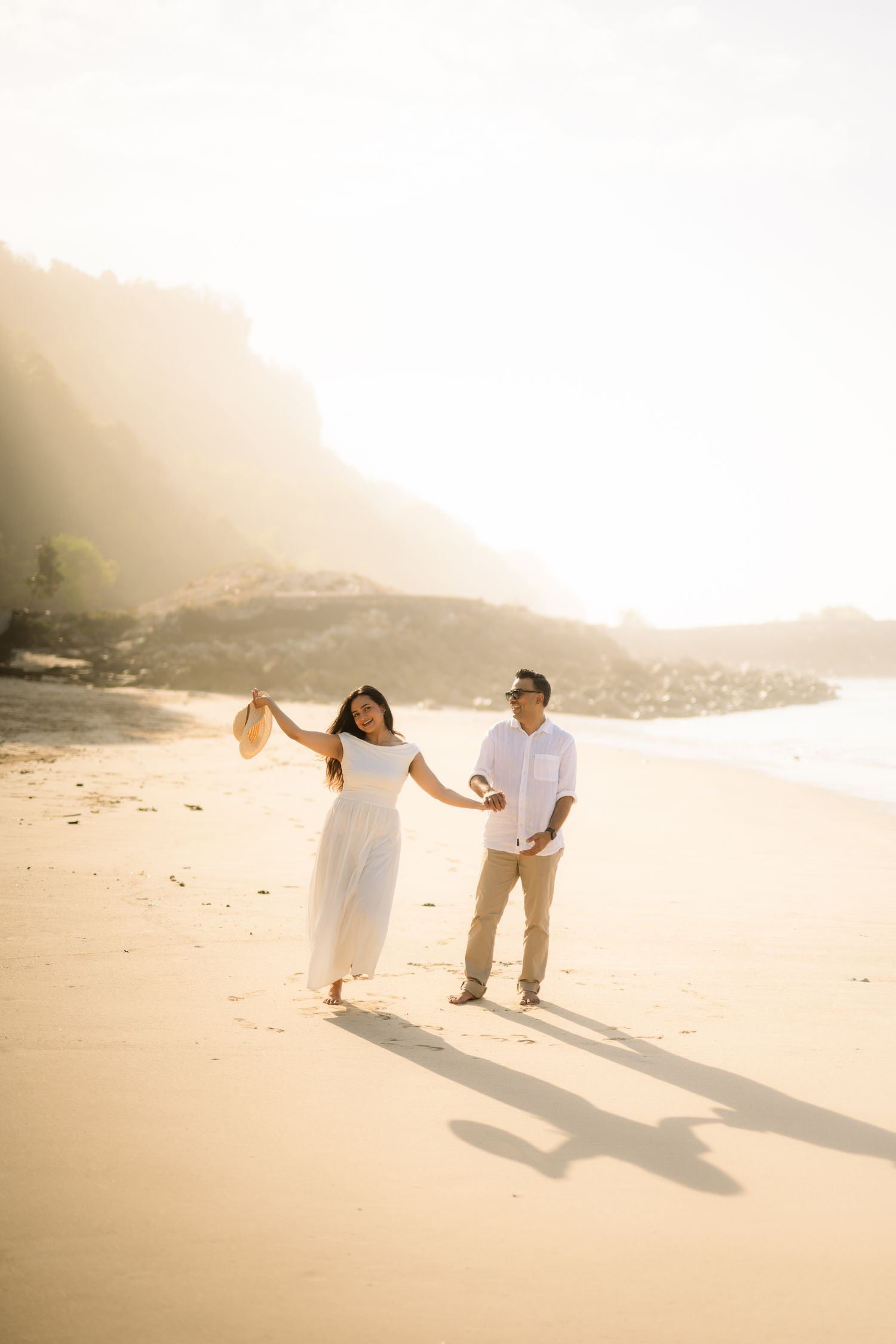 Couple walking hand in hand on a sunny beach, woman in white dress holding hat, man in white shirt and khaki pants.