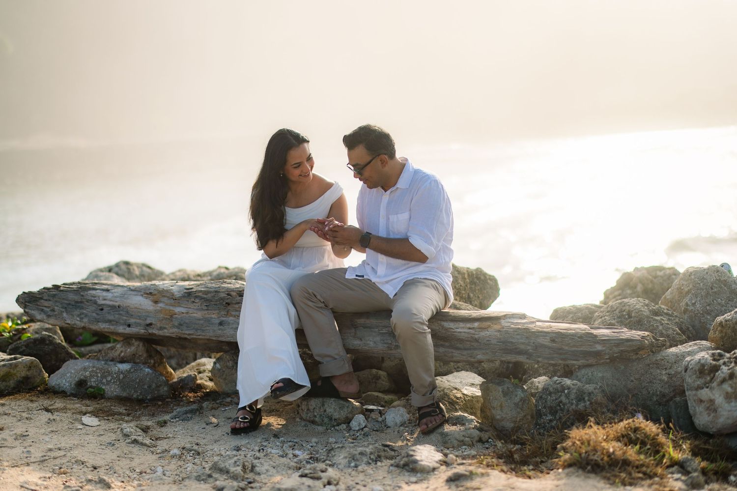 Couple sitting on driftwood at a sunny beach, sharing a romantic moment with ocean waves in the background.