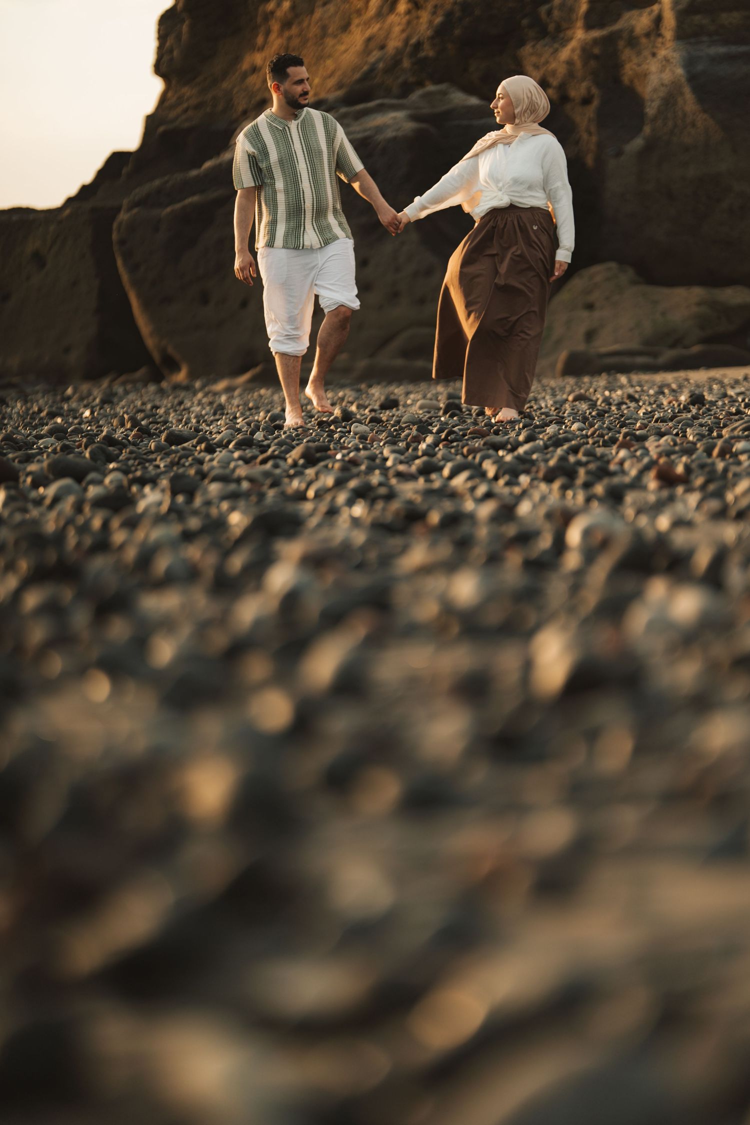 A couple holds hands while walking on a rocky beach at sunset with large boulders in the background.