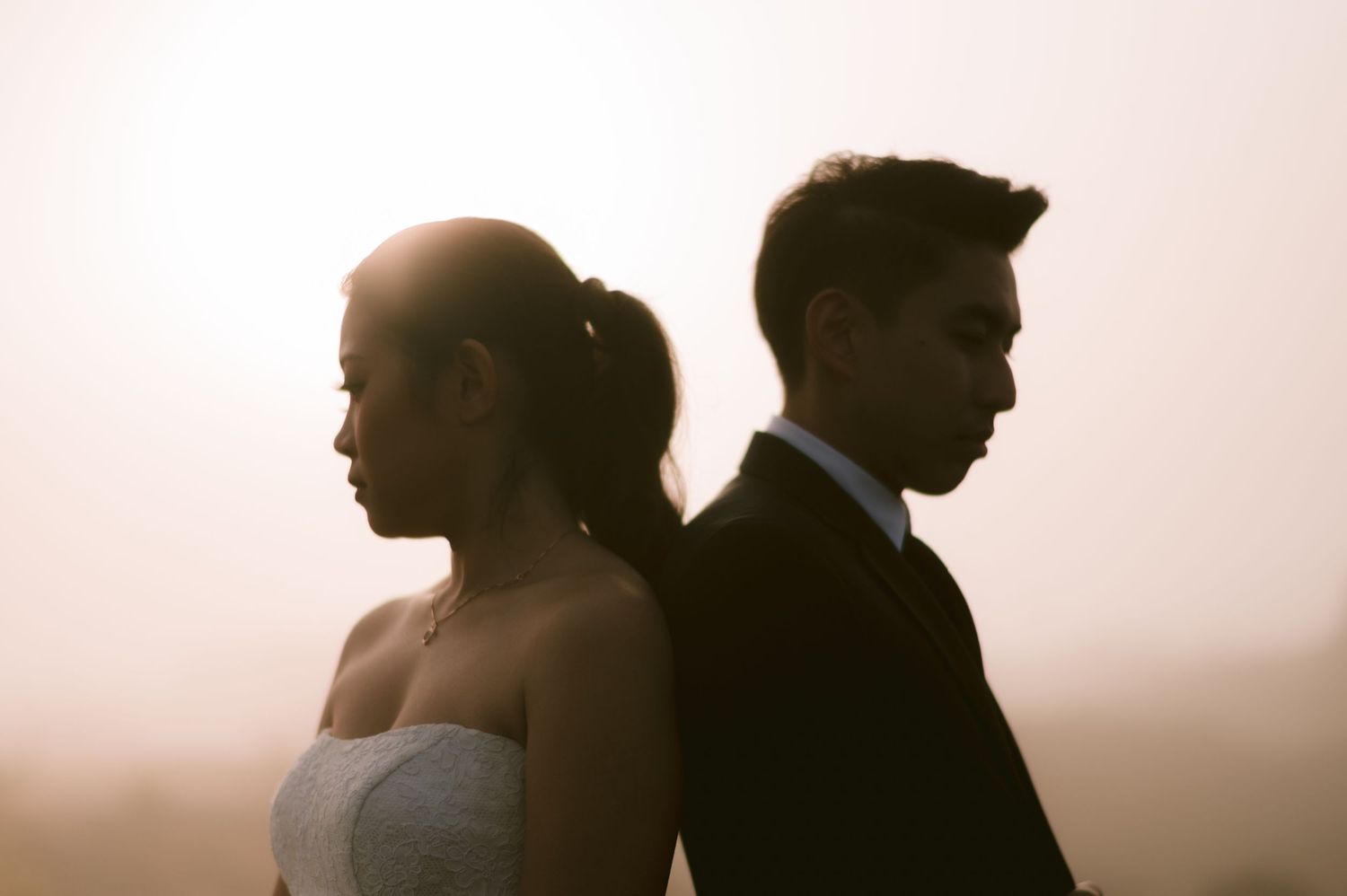 A bride and groom stand back-to-back in silhouette against a soft glowing sunset, symbolizing wedding tension.