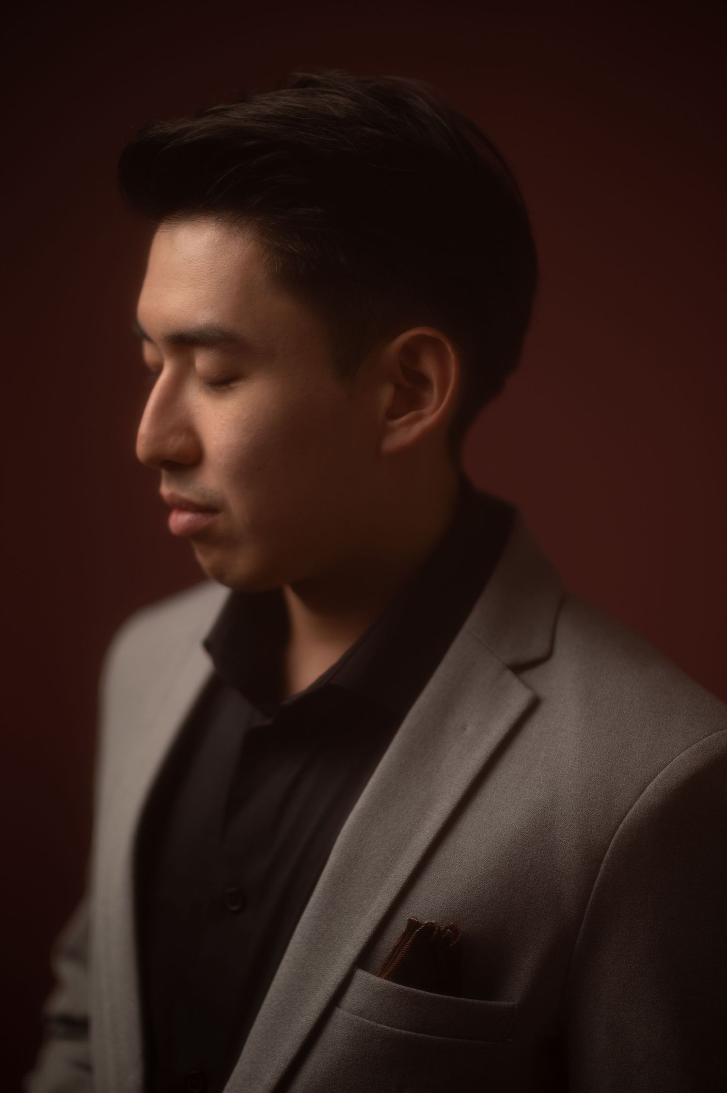 A young Asian man in a gray blazer and black shirt poses in profile with eyes closed against a dark red background.