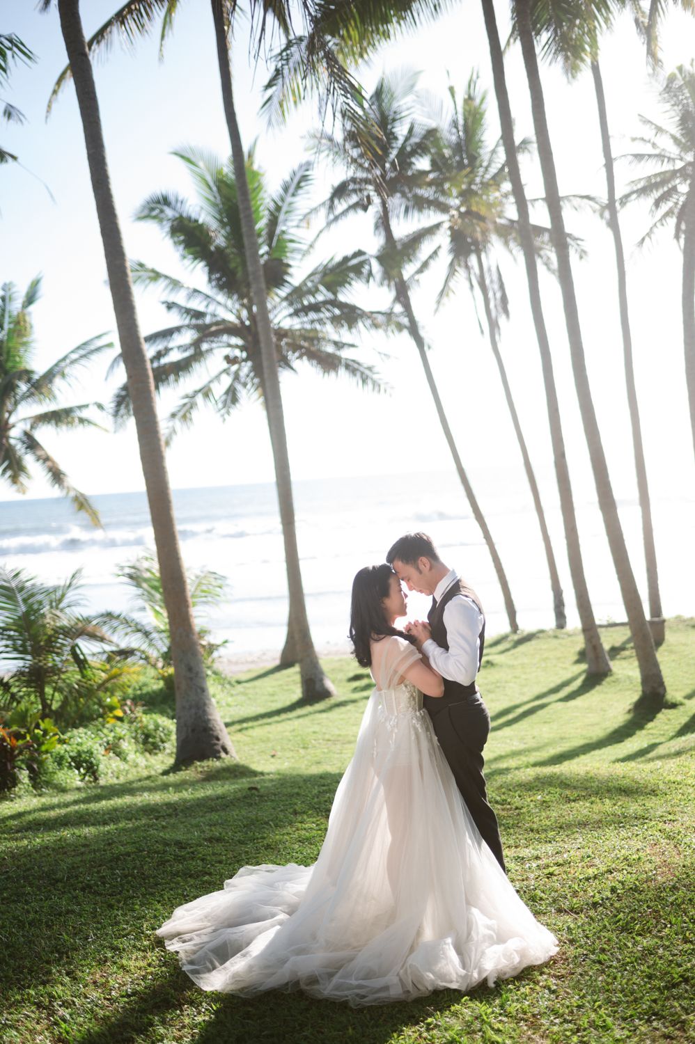 Bride and groom share a romantic kiss under tall palm trees on a lush green lawn with ocean views.