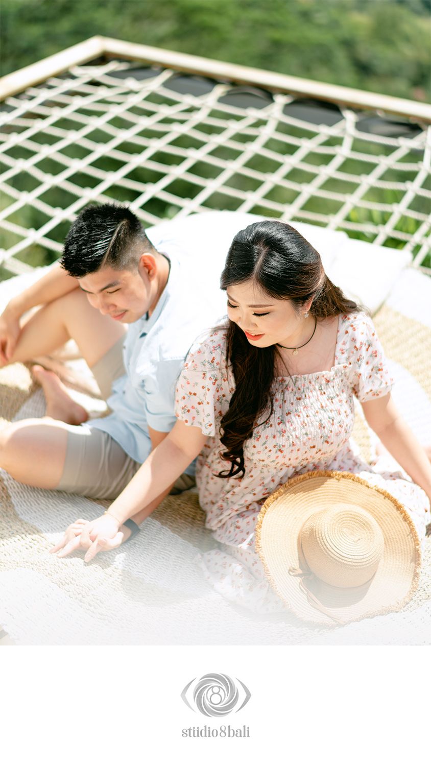 Couple sitting together on a woven mat, woman in floral dress with straw hat, man in light blue shirt, lush greenery backdrop.