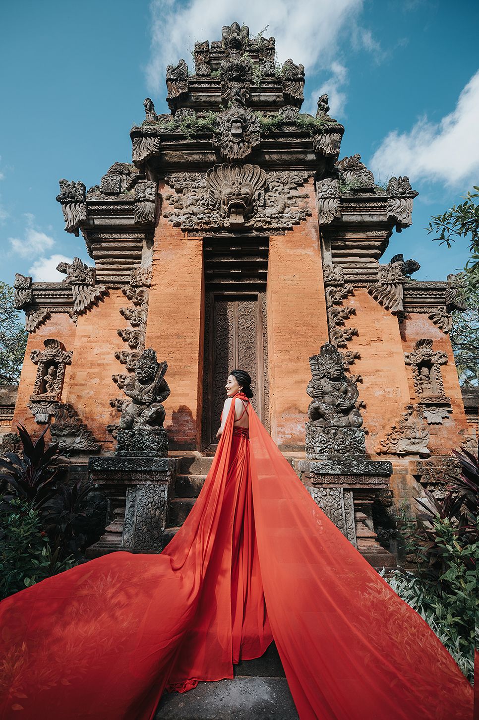 Woman in flowing red gown poses before ornate Balinese temple gate with dramatic fabric trailing behind her.