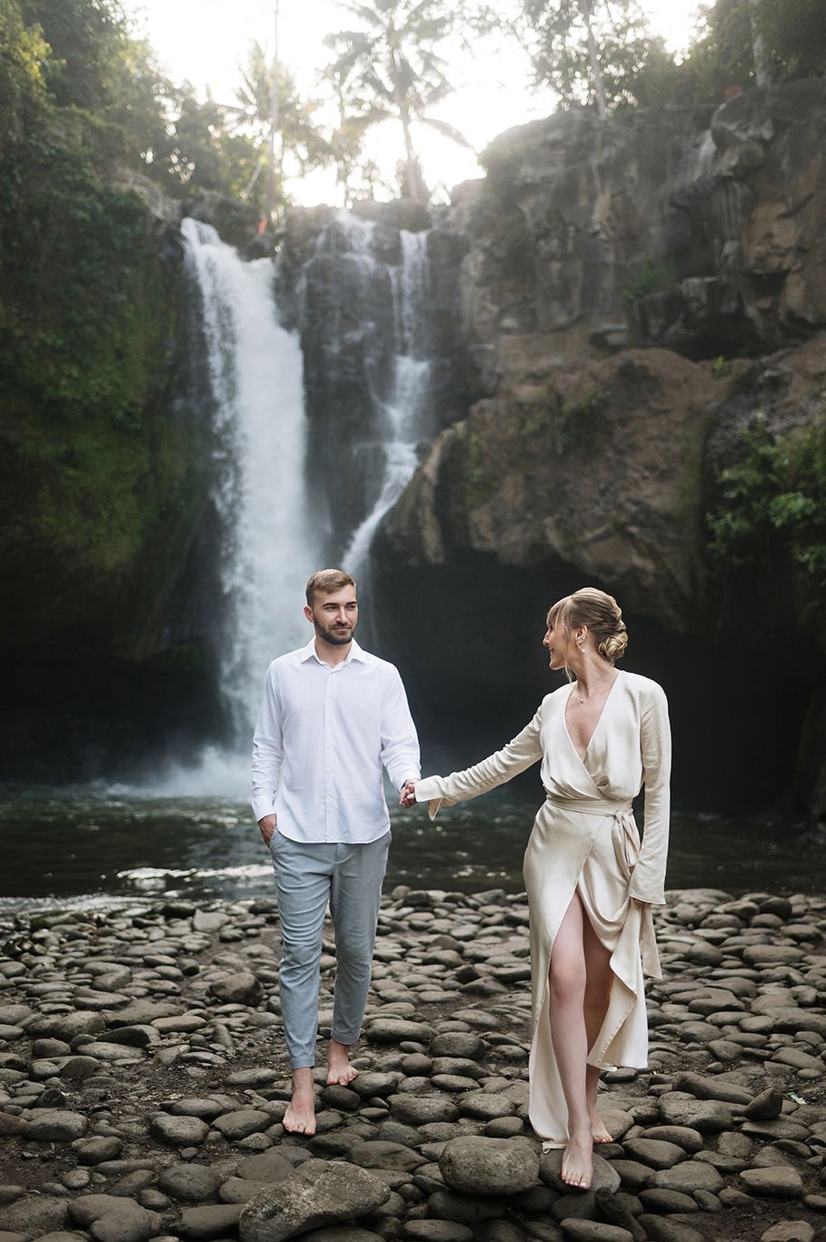 Couple holding hands barefoot on rocks before a tropical waterfall, man in white shirt and woman in elegant cream wrap dress.