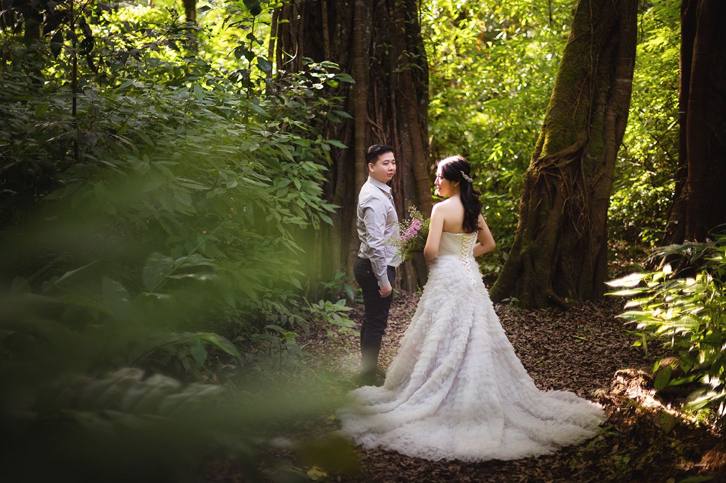 A bride and groom share a romantic moment on a forest path surrounded by lush green trees and ferns.