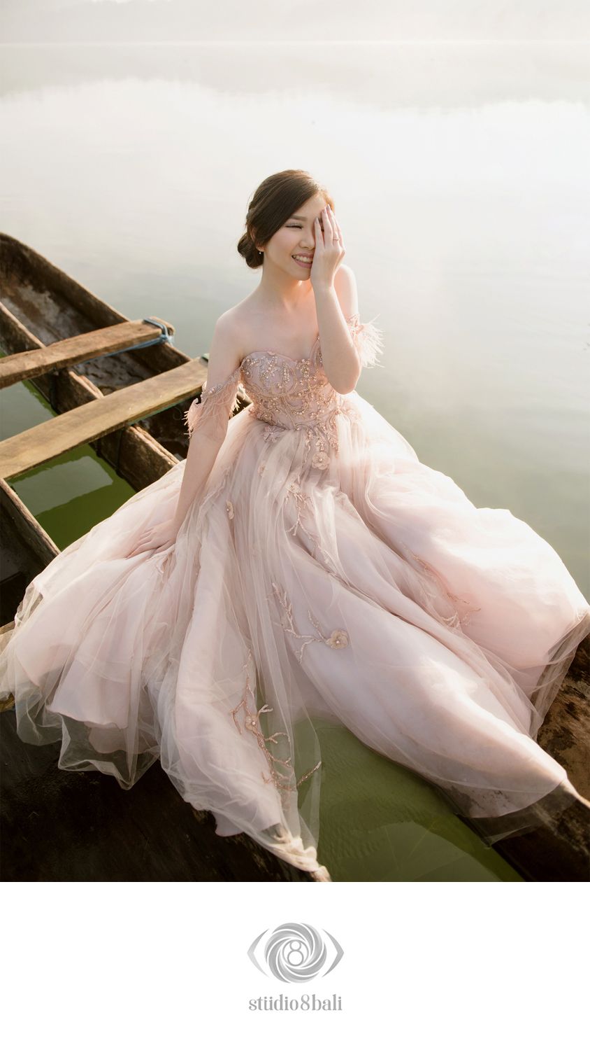 A bride in a blush pink ball gown sits gracefully in a wooden boat on calm water, laughing joyfully.
