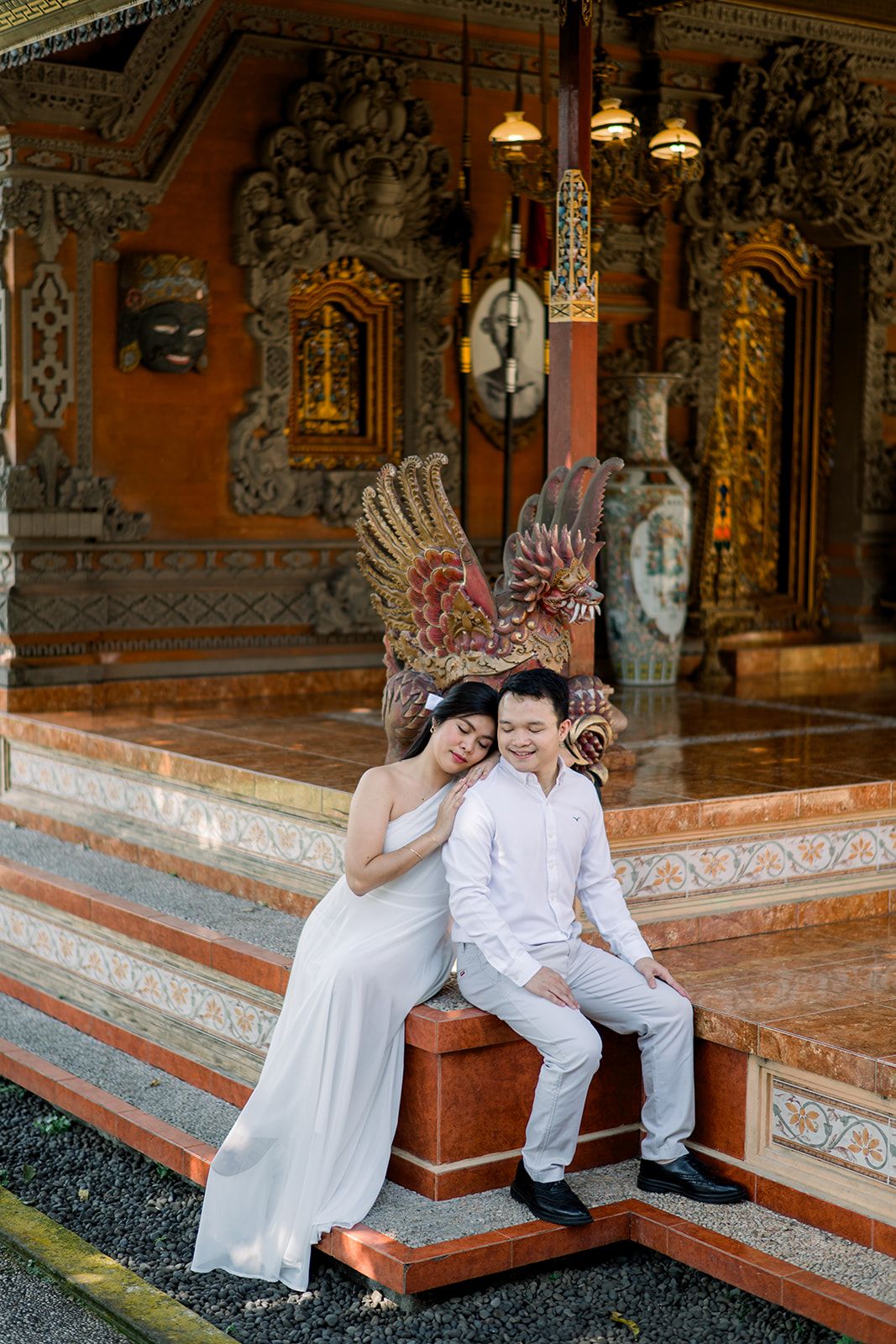 Couple in white attire posing romantically at an ornate Balinese temple with golden decorations.