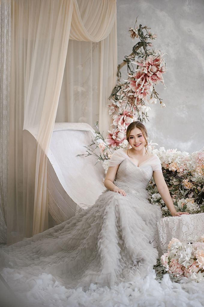Bride in stunning white feathered wedding gown sits among pink and white floral arrangements in dreamy studio.