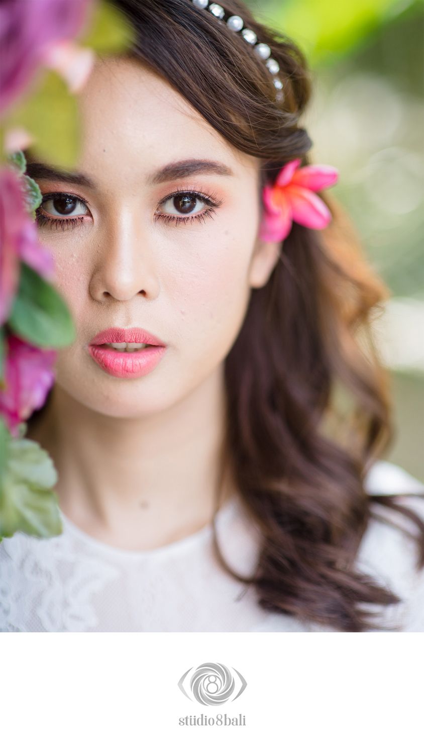 A beautiful Asian bride with floral hair accessories and pearl headband poses gracefully among pink tropical flowers.