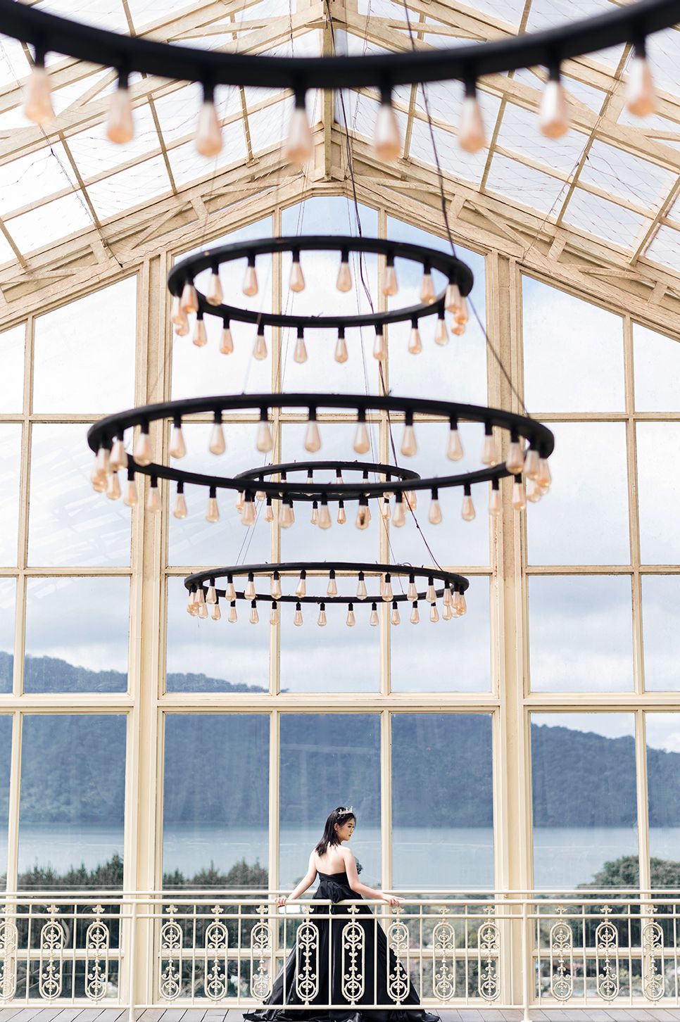 Woman in black gown stands in elegant glass greenhouse with tiered chandelier and scenic lake view.