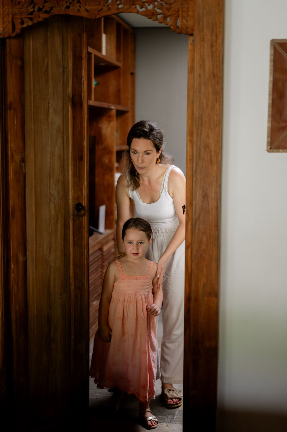 A mother and young daughter in a pink dress stand together in a wooden doorway in a warmly lit room.