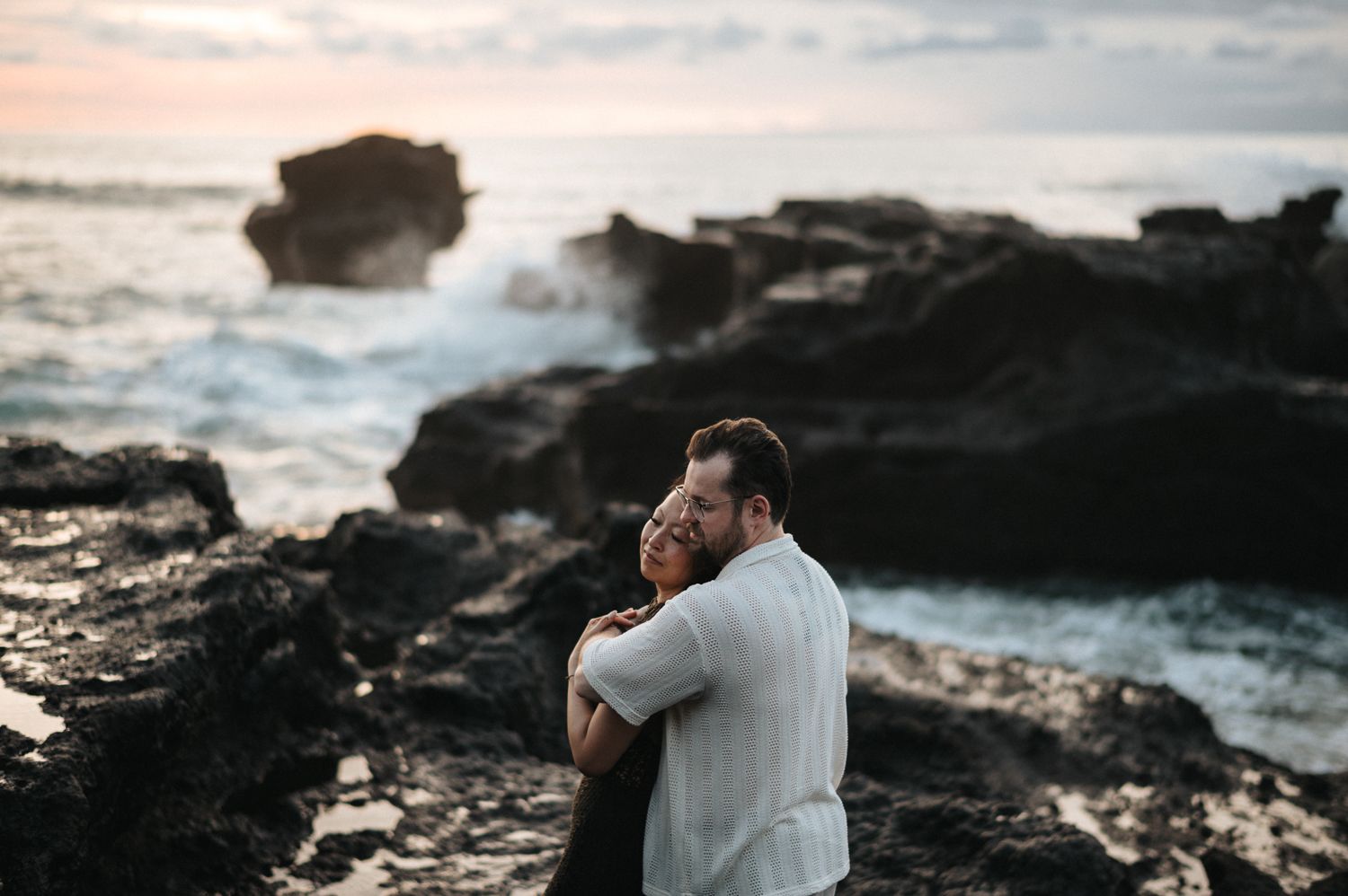 Couple embracing romantically on rocky ocean shore at sunset with crashing waves in background.