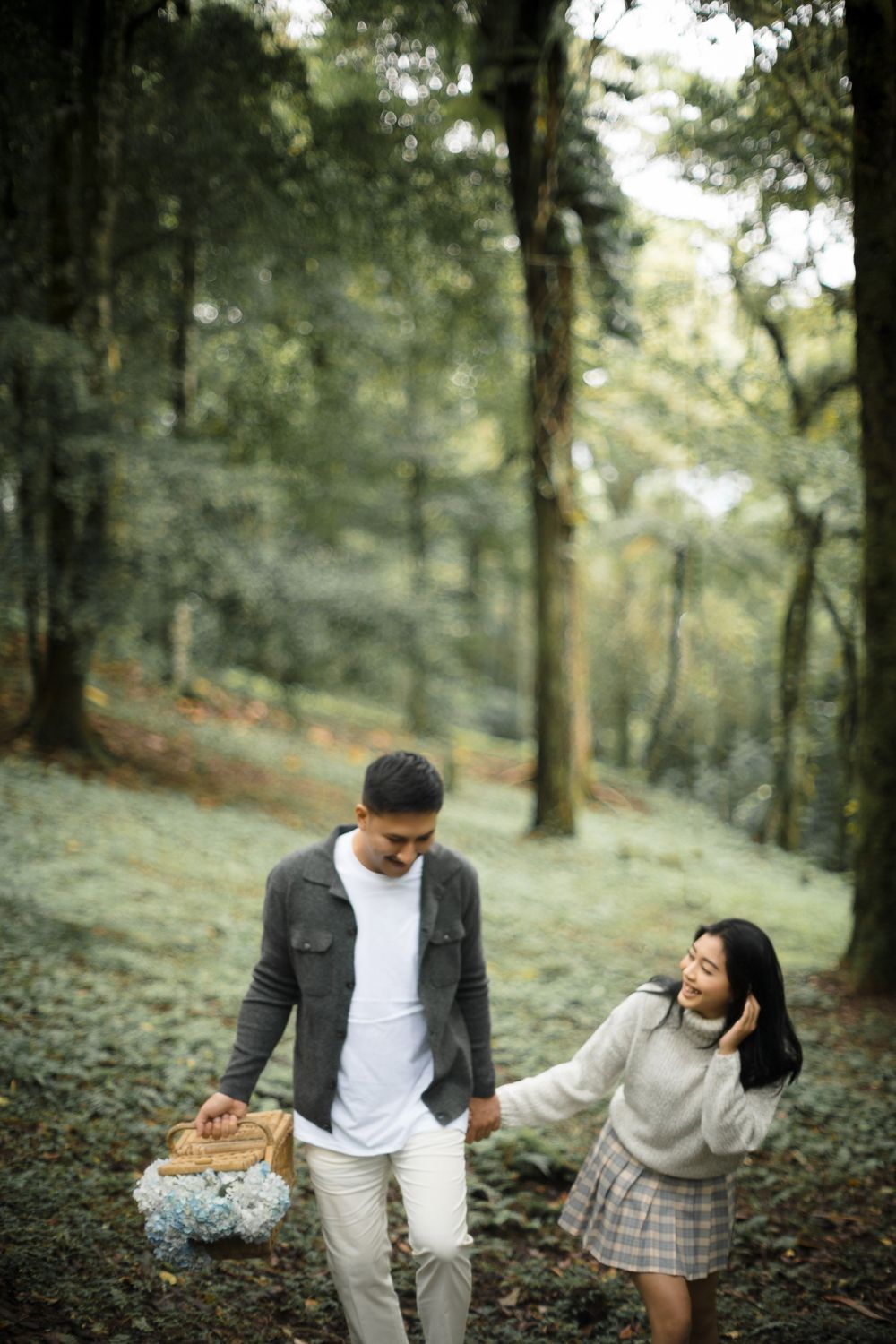 Couple walking hand-in-hand through a lush green forest, man carrying a picnic basket, woman smiling warmly at him.