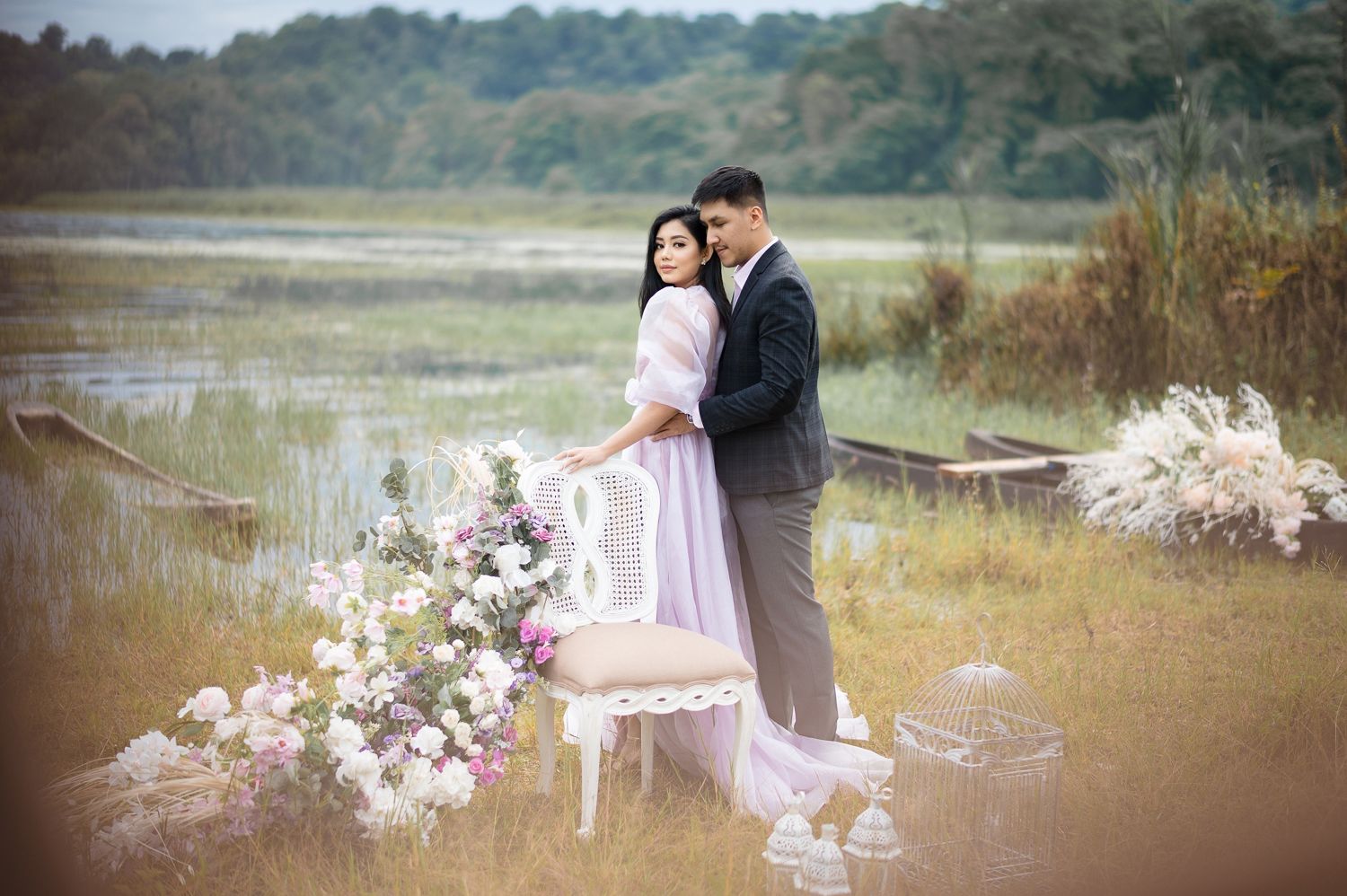Couple in elegant wedding attire embracing near a lake with floral arrangements and a vintage chair in a scenic outdoor setting.