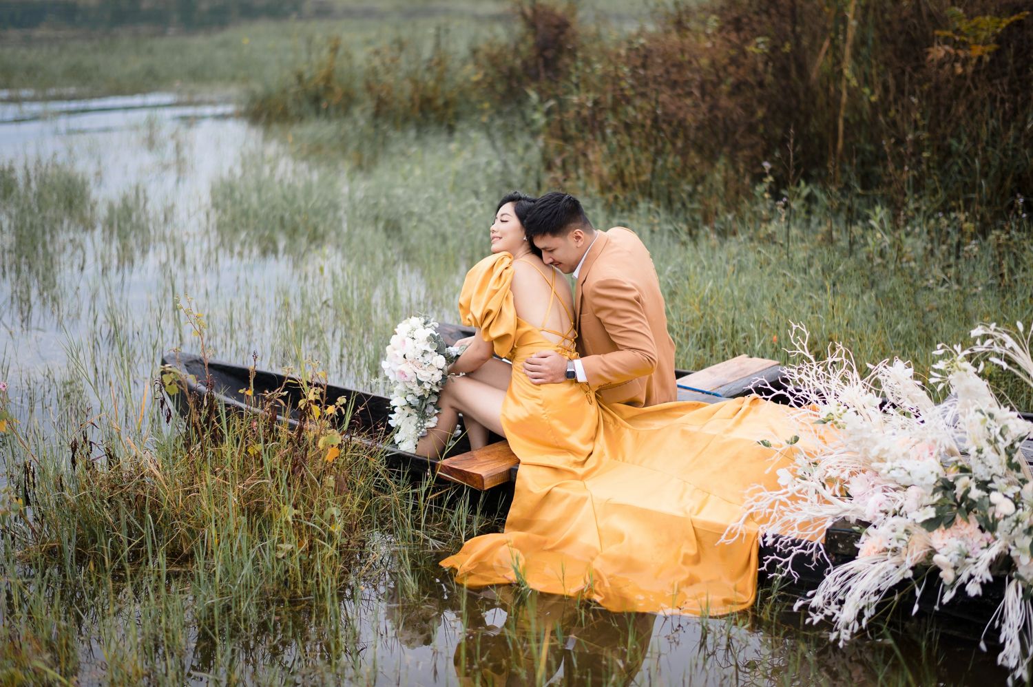 Couple in matching orange outfits sharing a romantic embrace in a rustic boat surrounded by wildflowers on a calm lake.