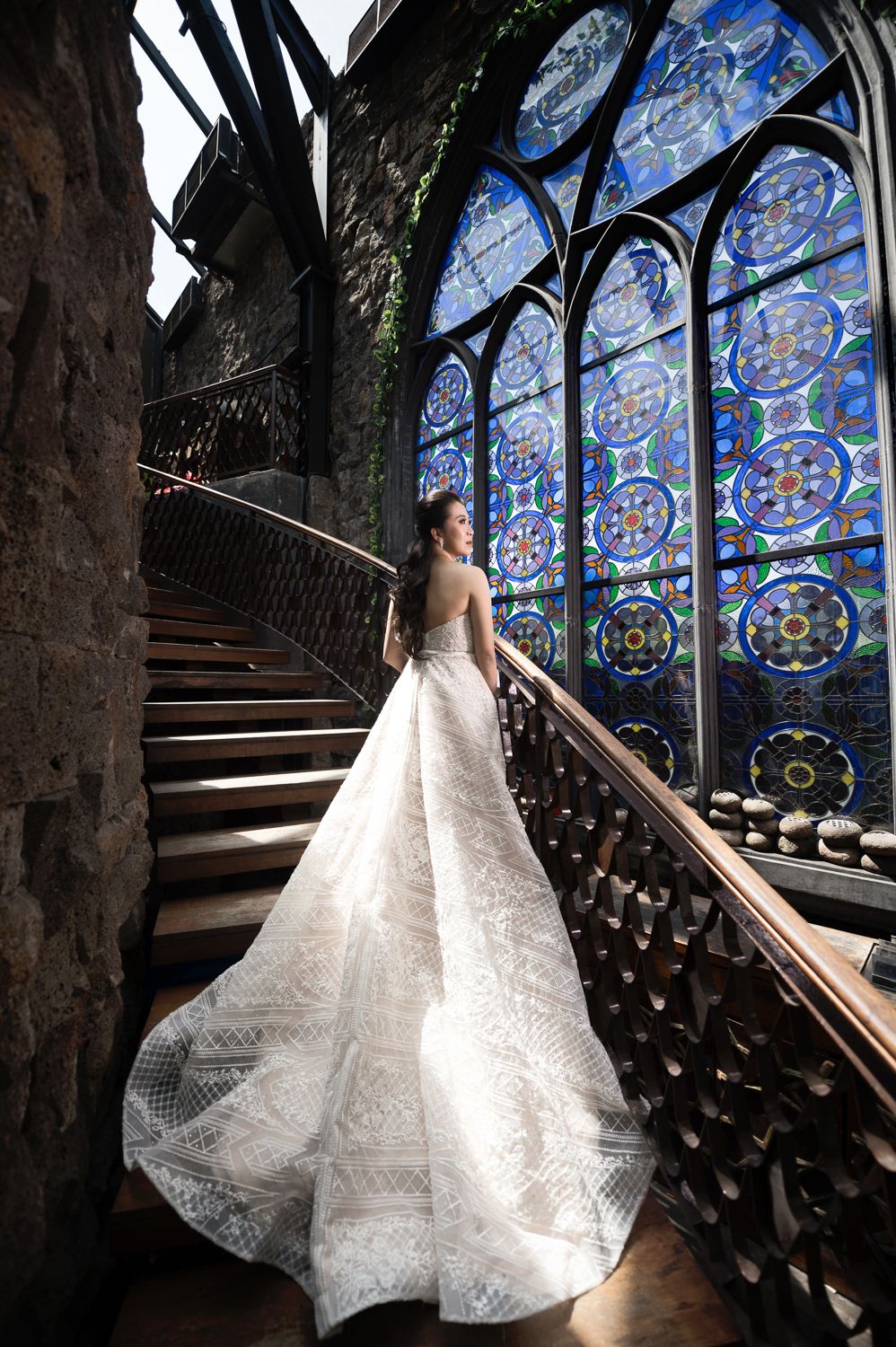 Bride in elegant lace gown with long train stands on ornate wooden staircase before stunning blue stained glass windows.