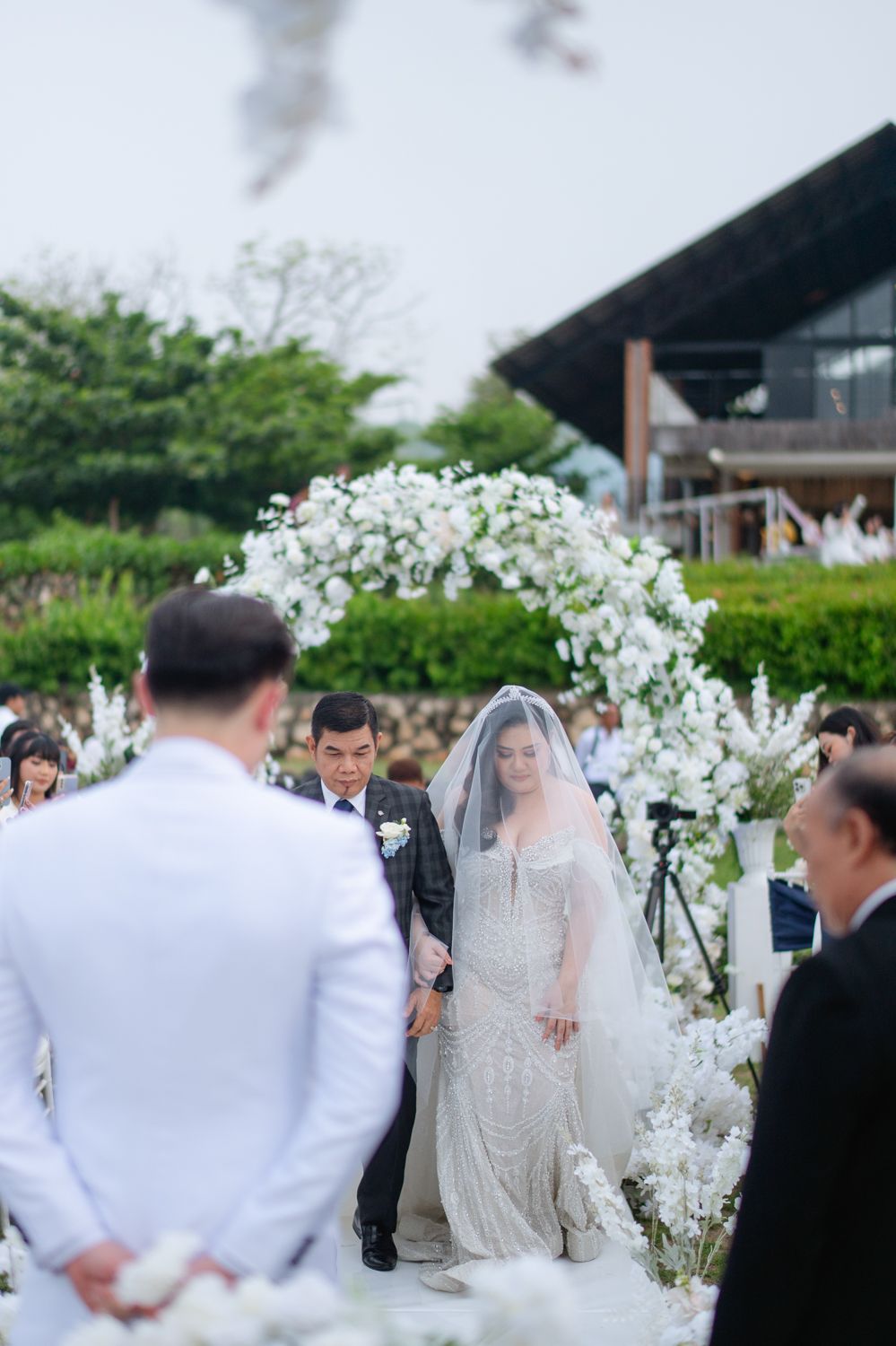 Bride in lace gown and veil walks down outdoor aisle under white floral arch toward groom in white suit.