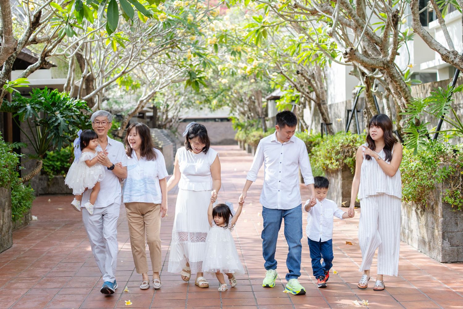 Multi-generational Asian family in white outfits walking together along a tree-lined garden pathway.