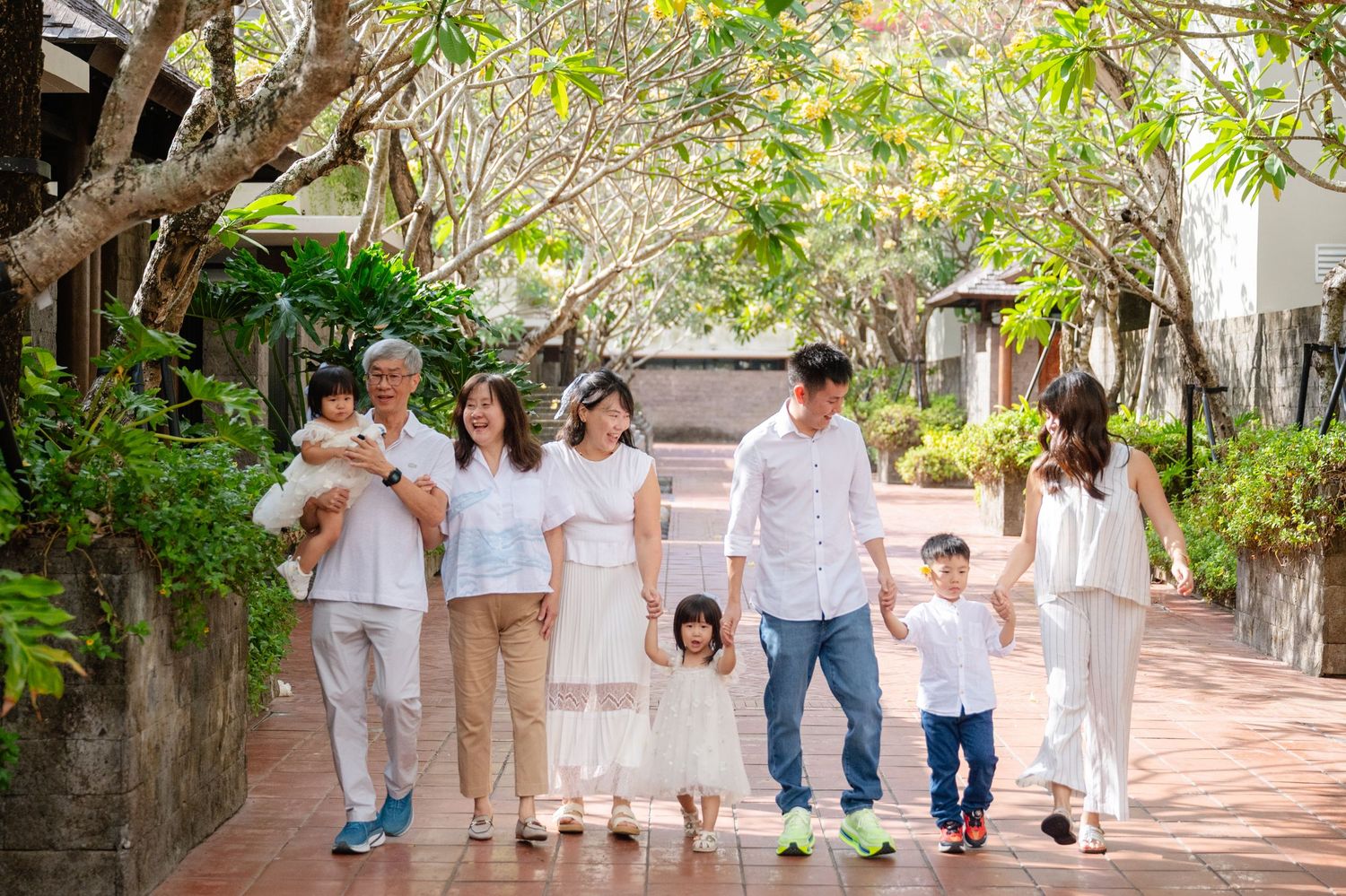 Multi-generational Asian family in white outfits walking together along a tree-lined path in a tropical garden.