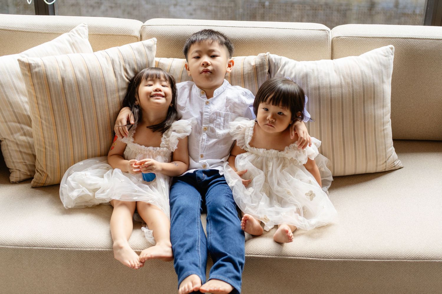 Three young Asian children sitting together on a beige sofa, two girls in white dresses and a boy in blue jeans.