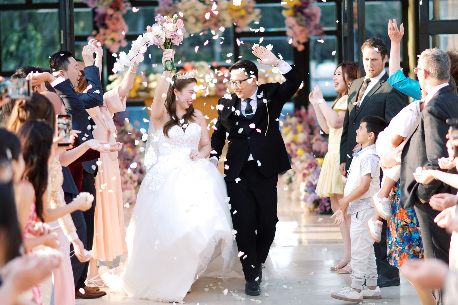 Joyful newlywed couple exits wedding ceremony as guests toss flower petals in celebration.
