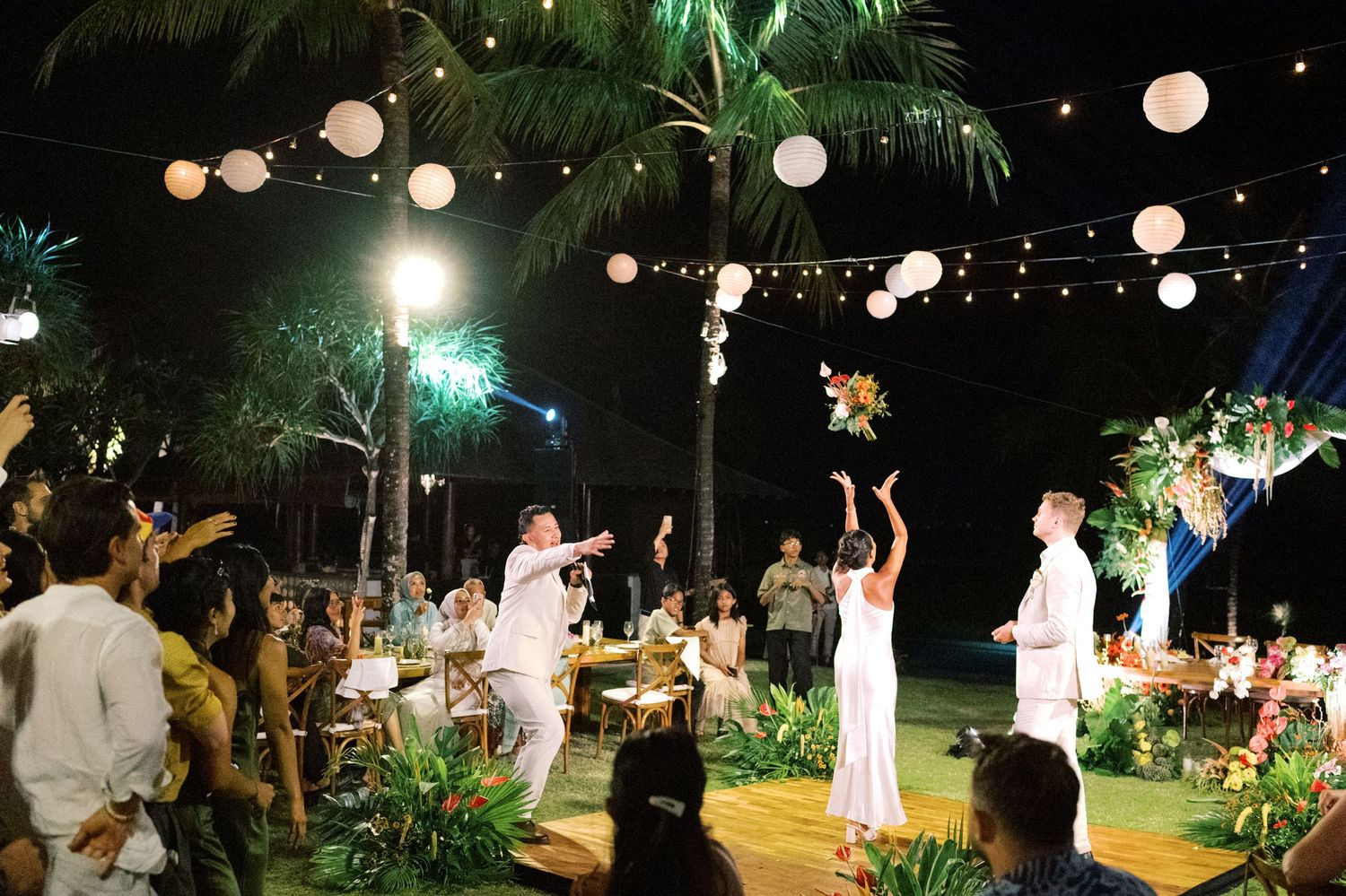 Outdoor tropical wedding reception at night with string lights, palm trees, and guests in white attire celebrating.