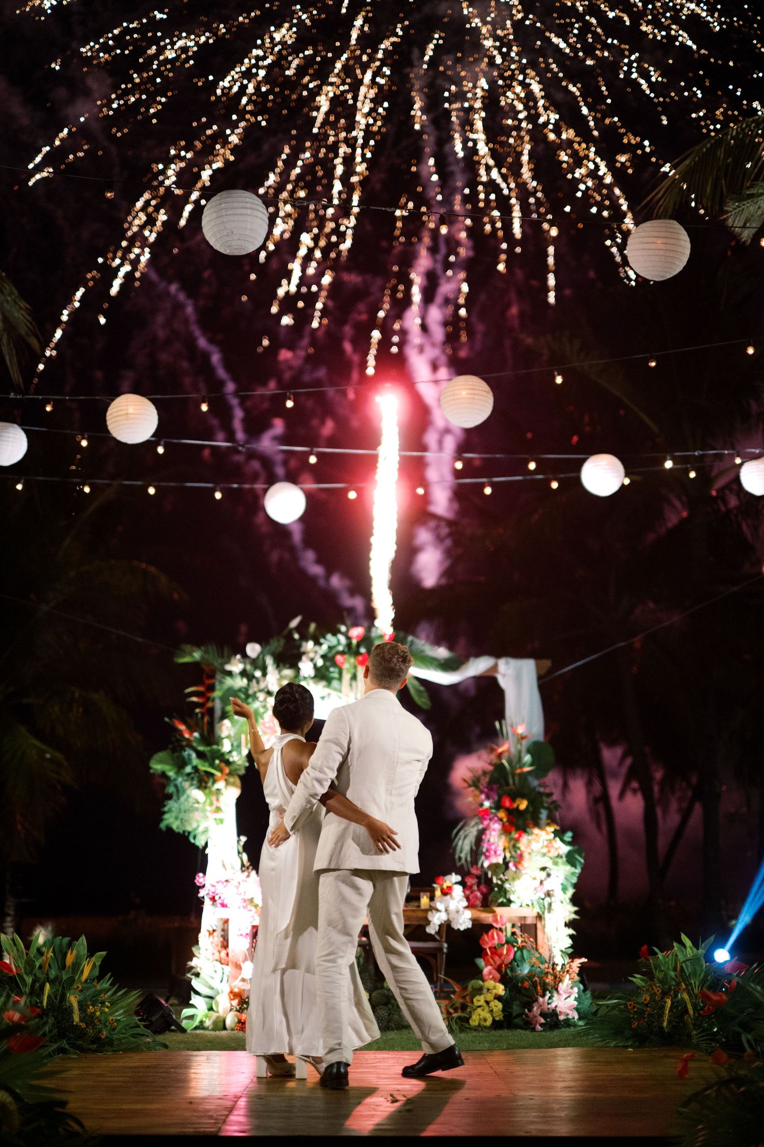 Couple in white dancing under fireworks and string lights with paper lanterns at an outdoor wedding reception.