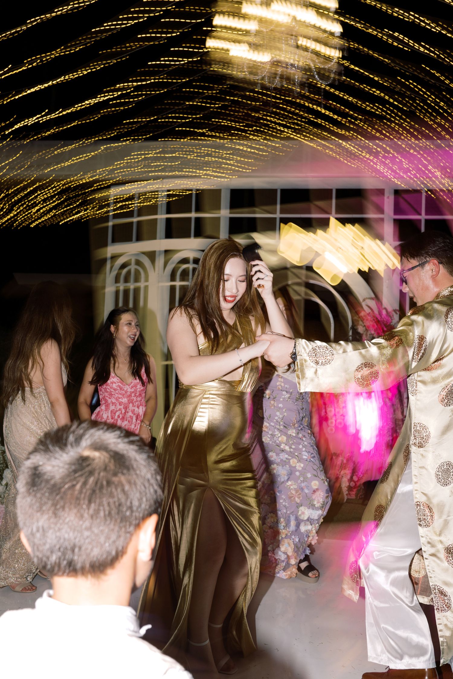 Guests dancing at a festive evening party with golden light trails and colorful pink lighting in the background.