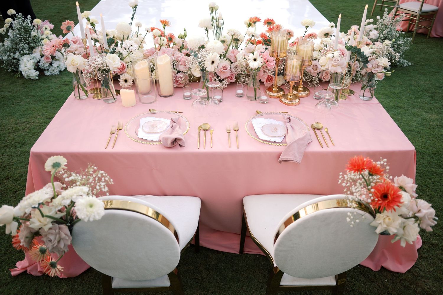 Elegant outdoor wedding sweetheart table with pink tablecloth, gold accents, floral arrangements, and white shell-shaped chairs.