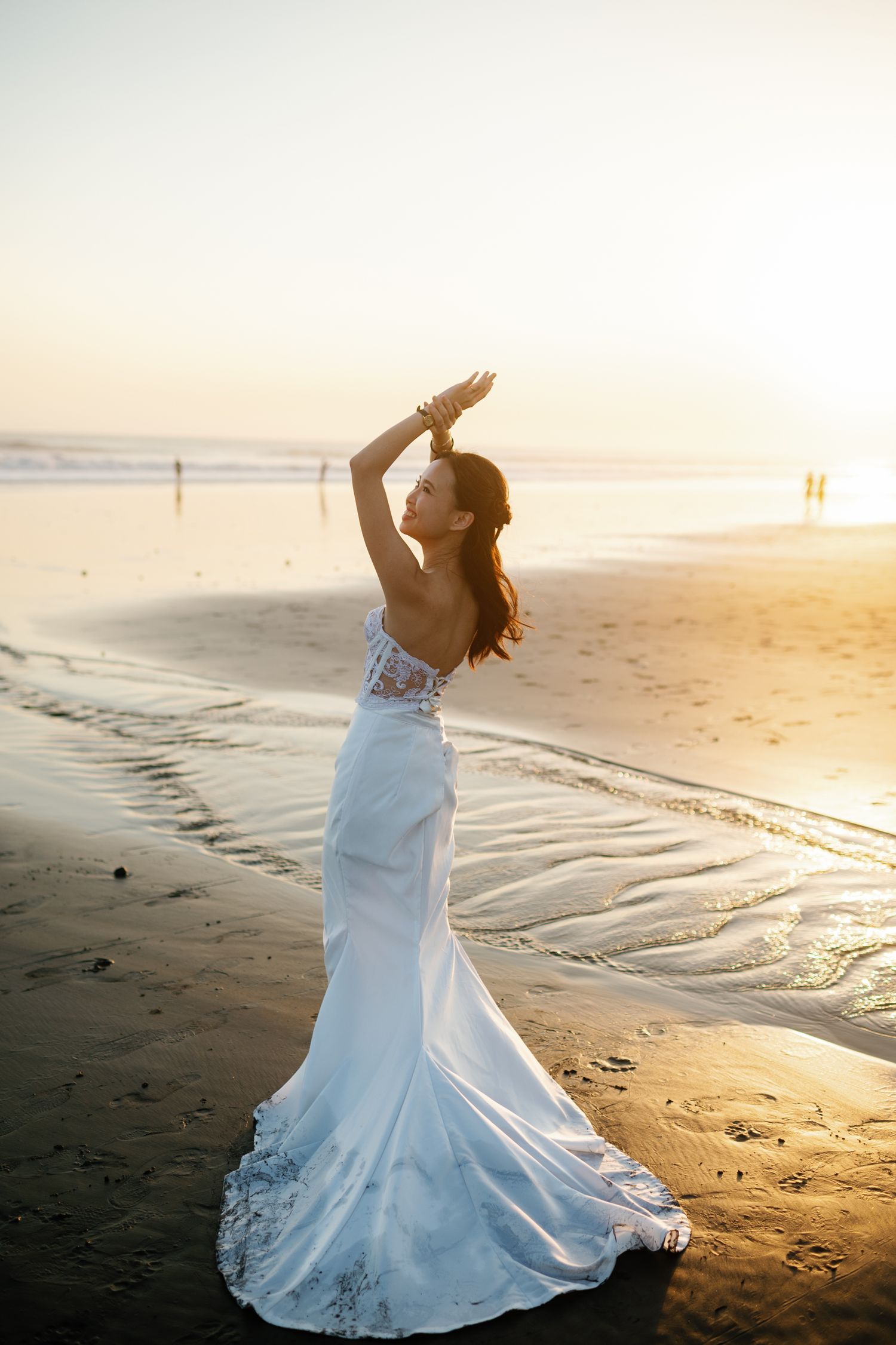 Bride in elegant white mermaid gown posing gracefully on a sandy beach at golden sunset.