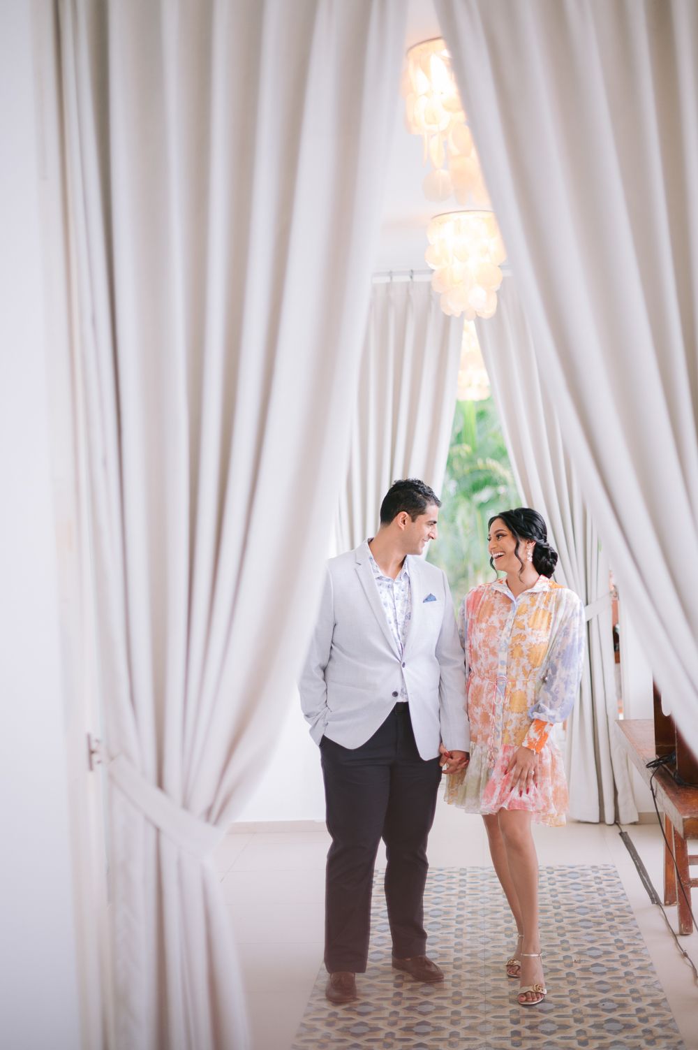 Couple holding hands and smiling at each other in an elegant white draped hallway with chandelier lighting.