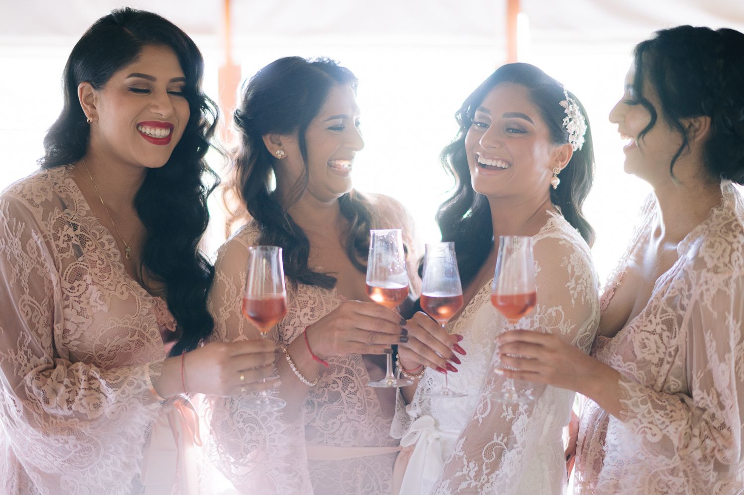 Four bridesmaids in blush lace dresses smiling and toasting with rose wine glasses at a wedding celebration.