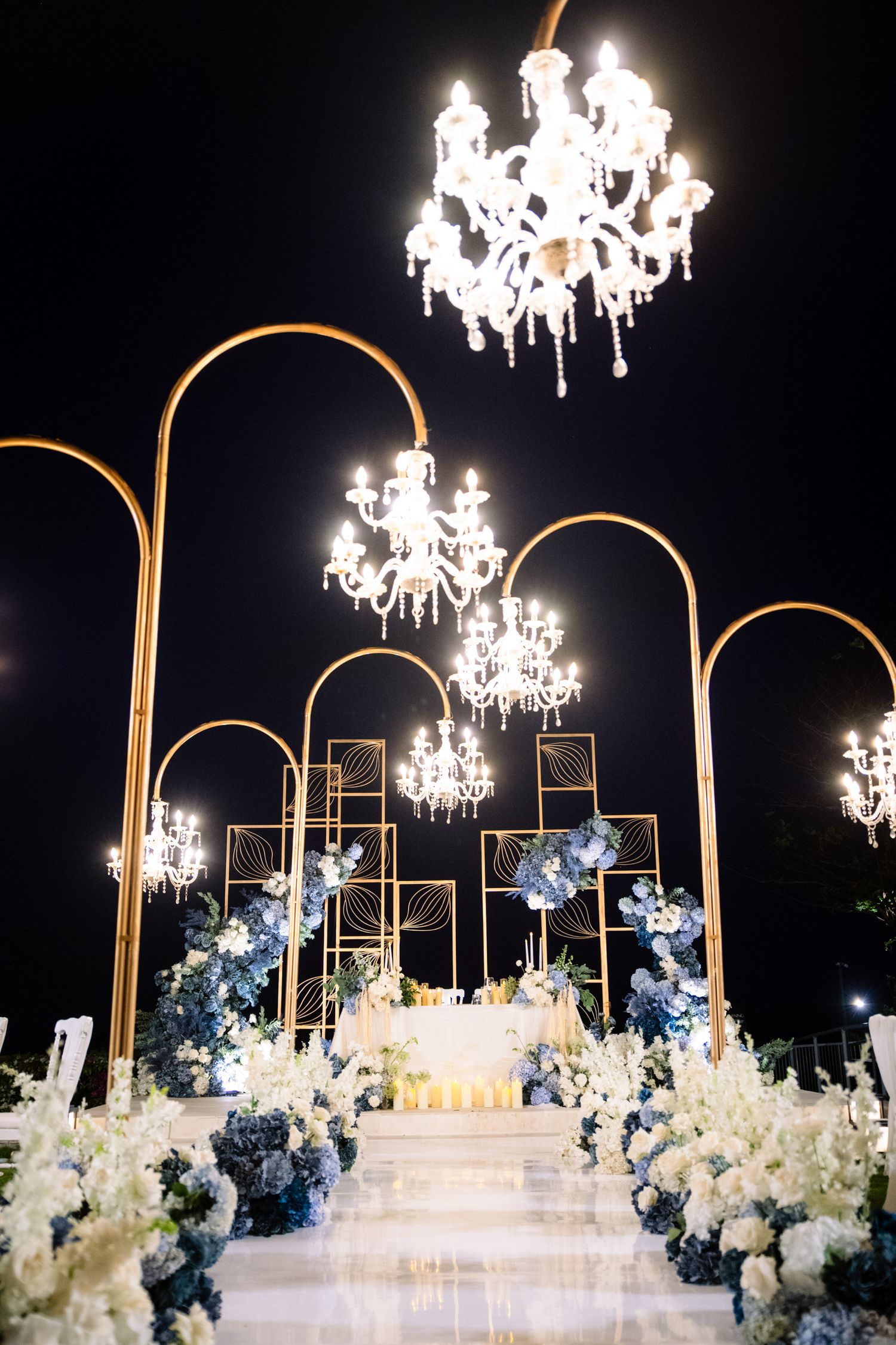 Elegant outdoor night wedding ceremony with gold arches, crystal chandeliers, and blue and white floral arrangements lining a glowing aisle.