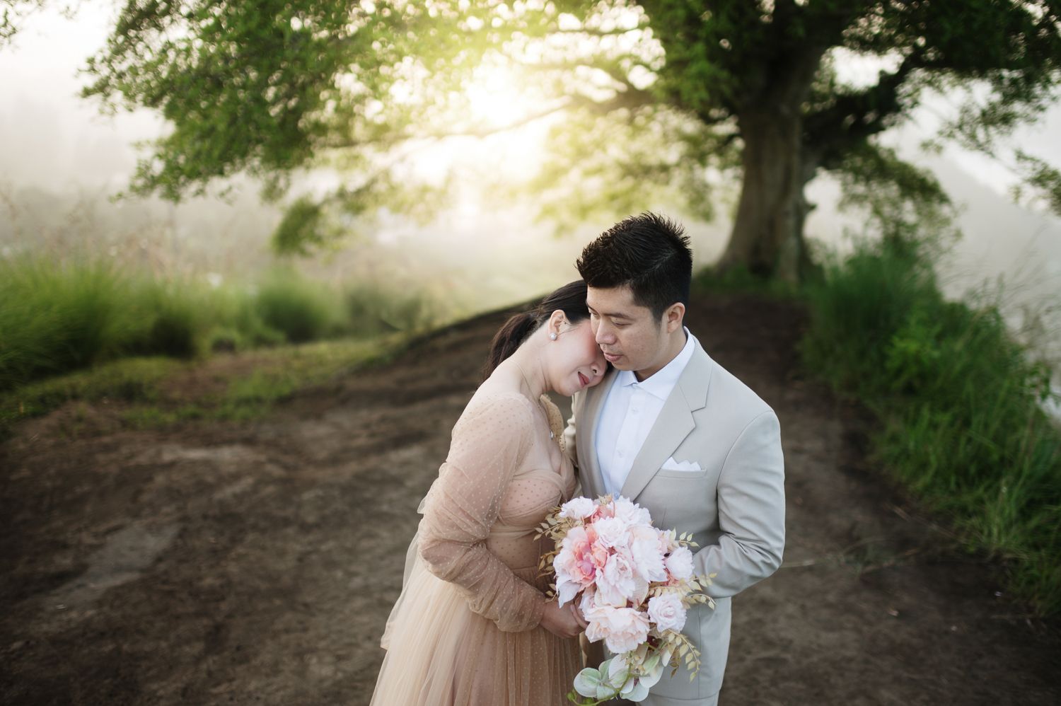 Romantic couple embracing outdoors near a misty tree, bride in pink gown holding bouquet, groom in gray suit.
