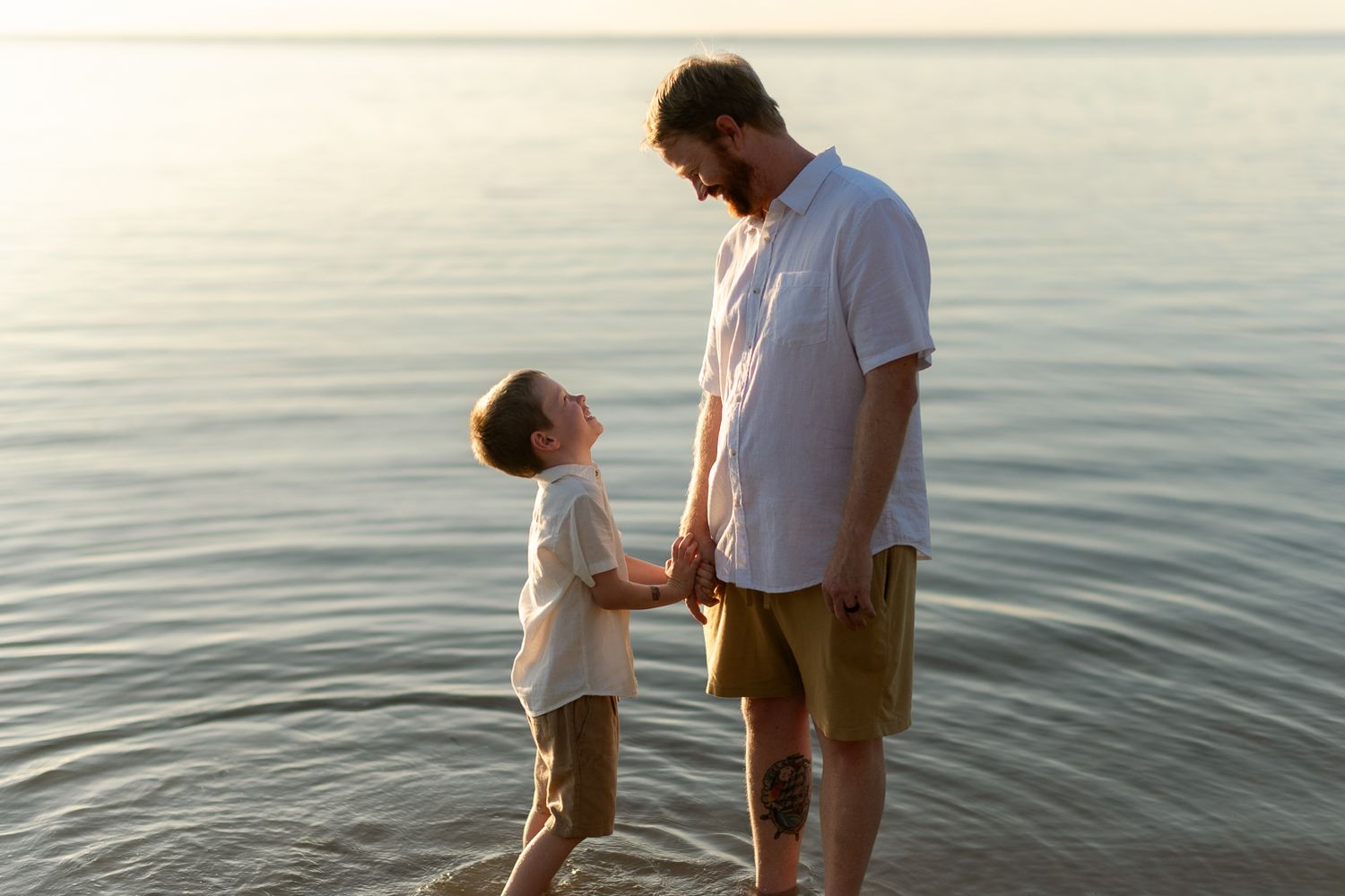 Father and young son holding hands while standing in calm ocean water at golden sunset.
