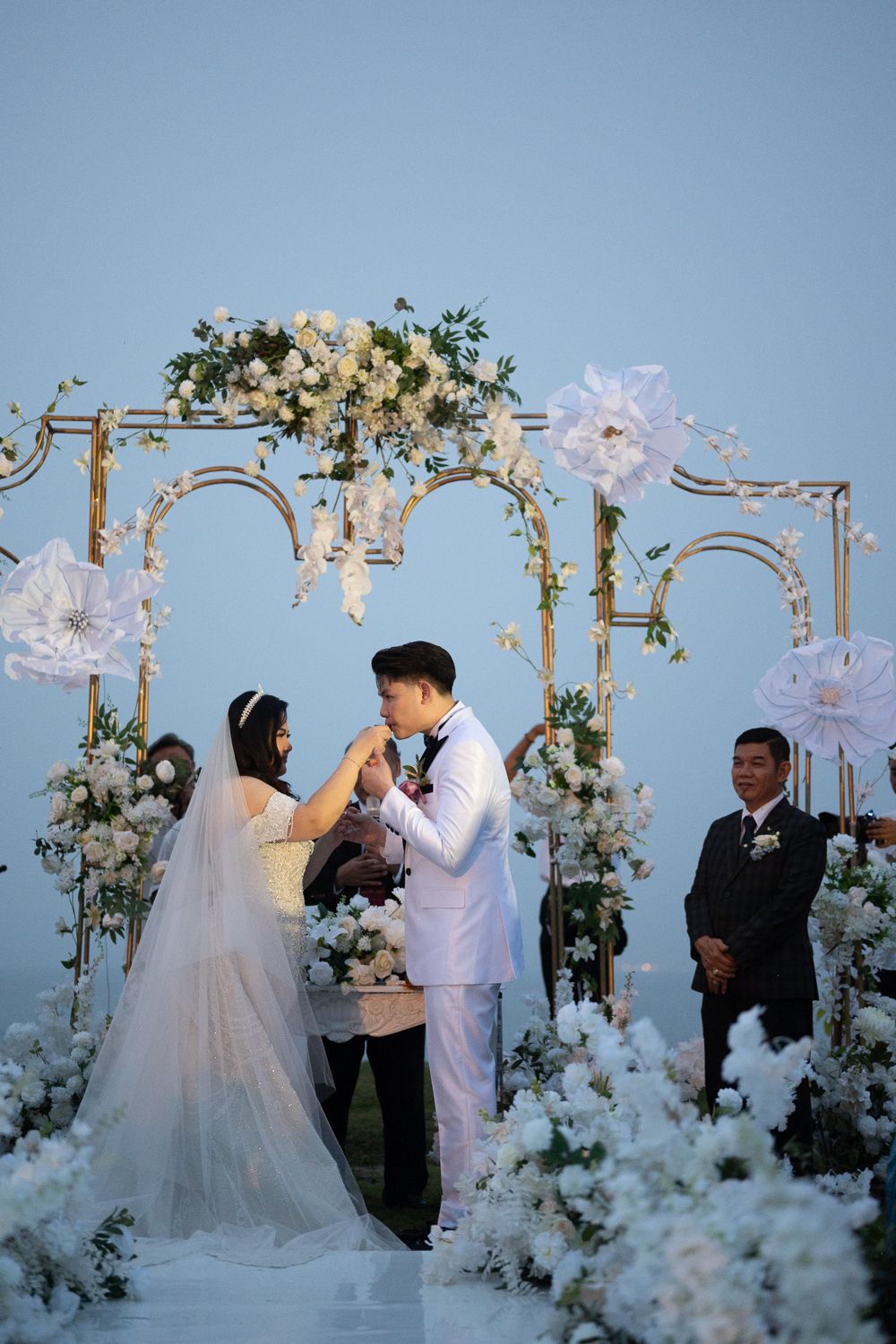 Couple exchanging vows at an outdoor wedding ceremony beneath a floral arch adorned with white roses and paper flowers.