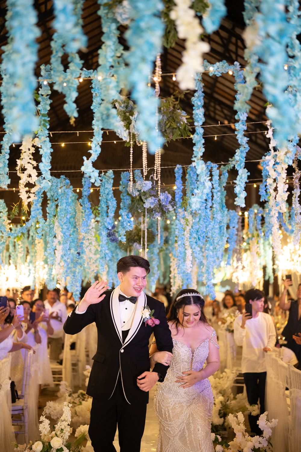 Newlywed couple walks down the aisle under stunning blue floral ceiling decorations at elegant wedding reception.