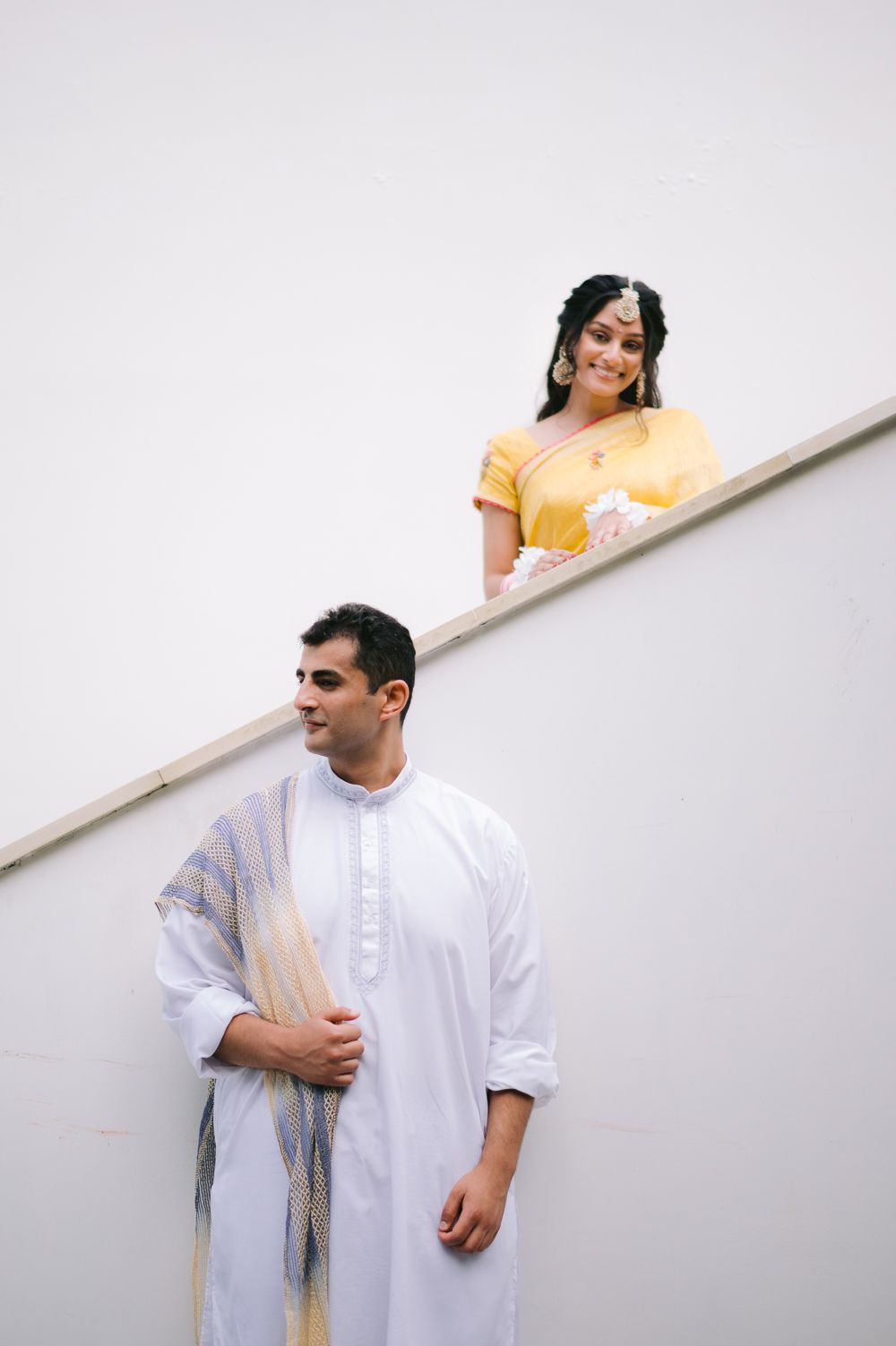 Indian couple in traditional attire, man in white kurta and woman in yellow lehenga smiling on staircase.